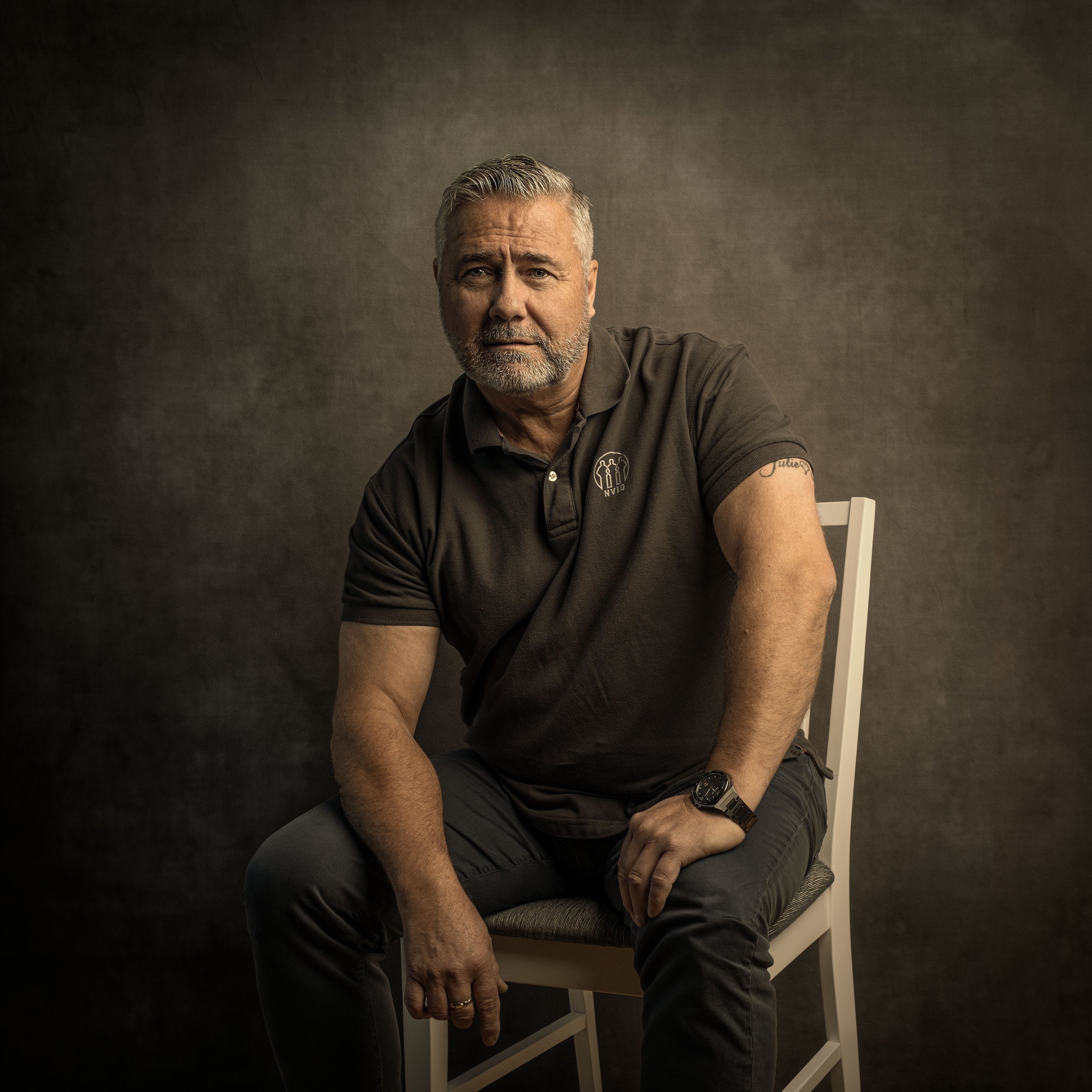 Et portrett av en eldre mann med grått hår og skjegg, som sitter på en stol mot en mørk bakgrunn, iført en svart polo med en logo og har armbåndsur på venstre hånd.