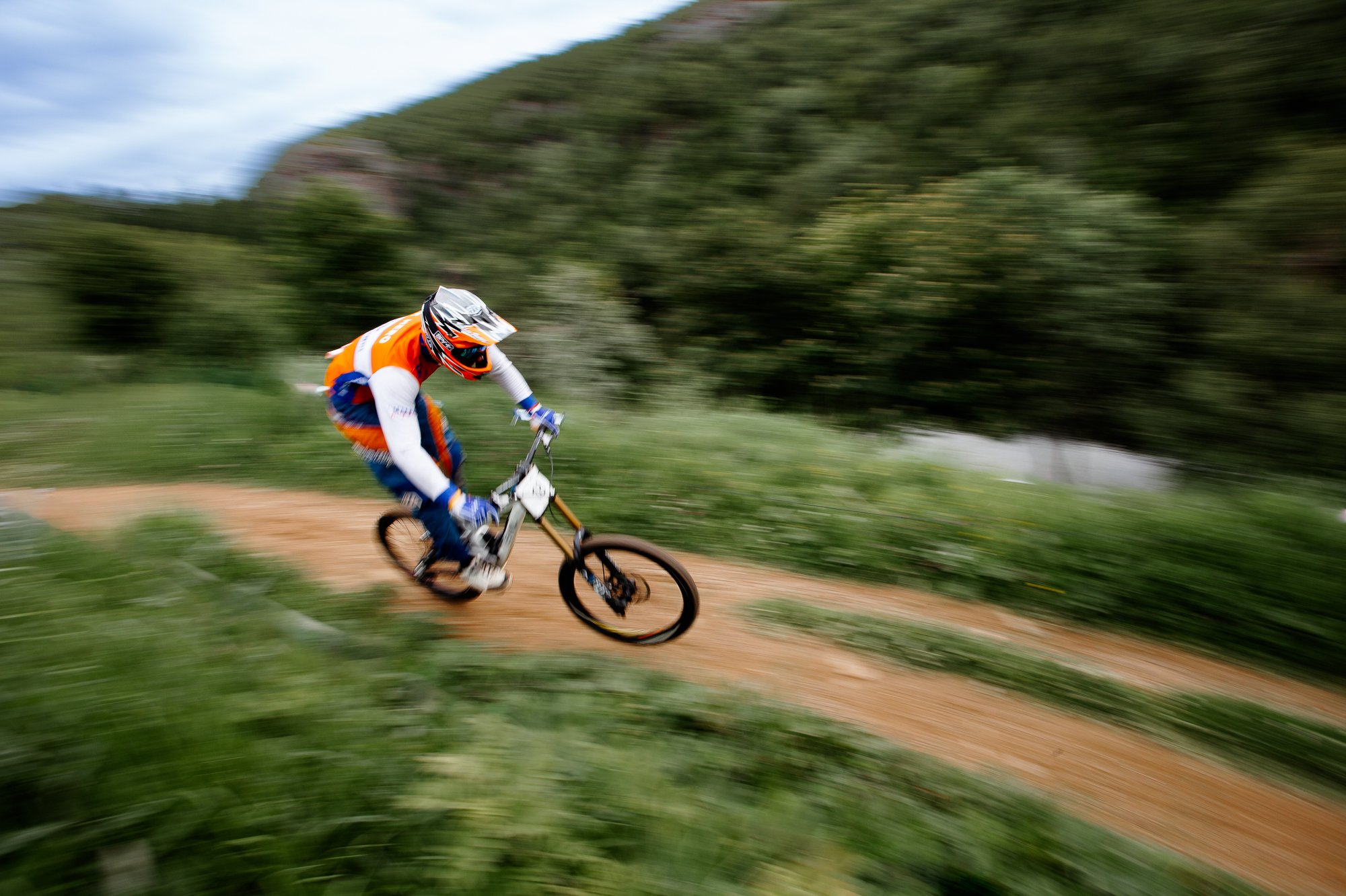 En syklist kjører raskt på en ugruset sykkelvei i naturen, omgitt av grønne planter og bakke. Syklisten er iført hjelm og fargerikt utstyr og sykler på en terrengsykkel.