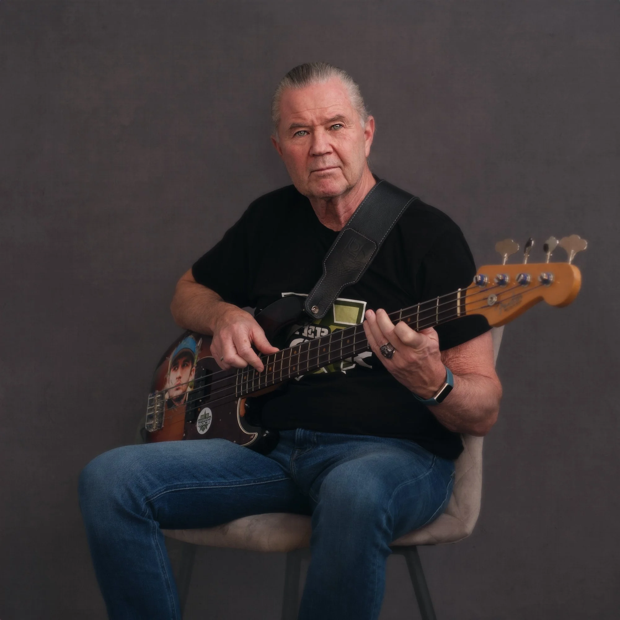 En eldre mann med grått hår som spiller bassgitar, sitter på en stol mot en grå bakgrunn.