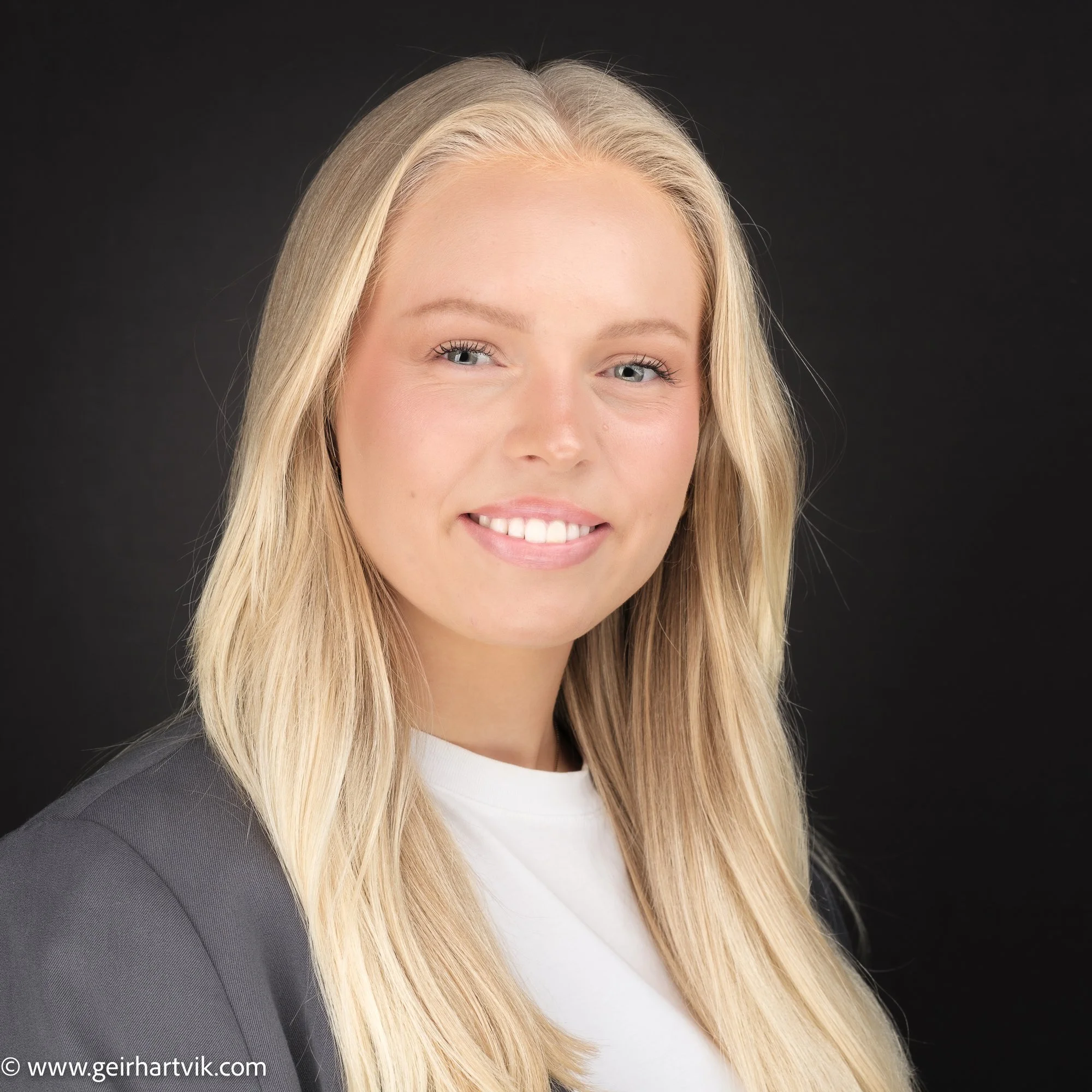 Porträtt av en ung kvinna med långt blont hår, leende, bär svart kavaj och vit topp mot svart bakgrund.