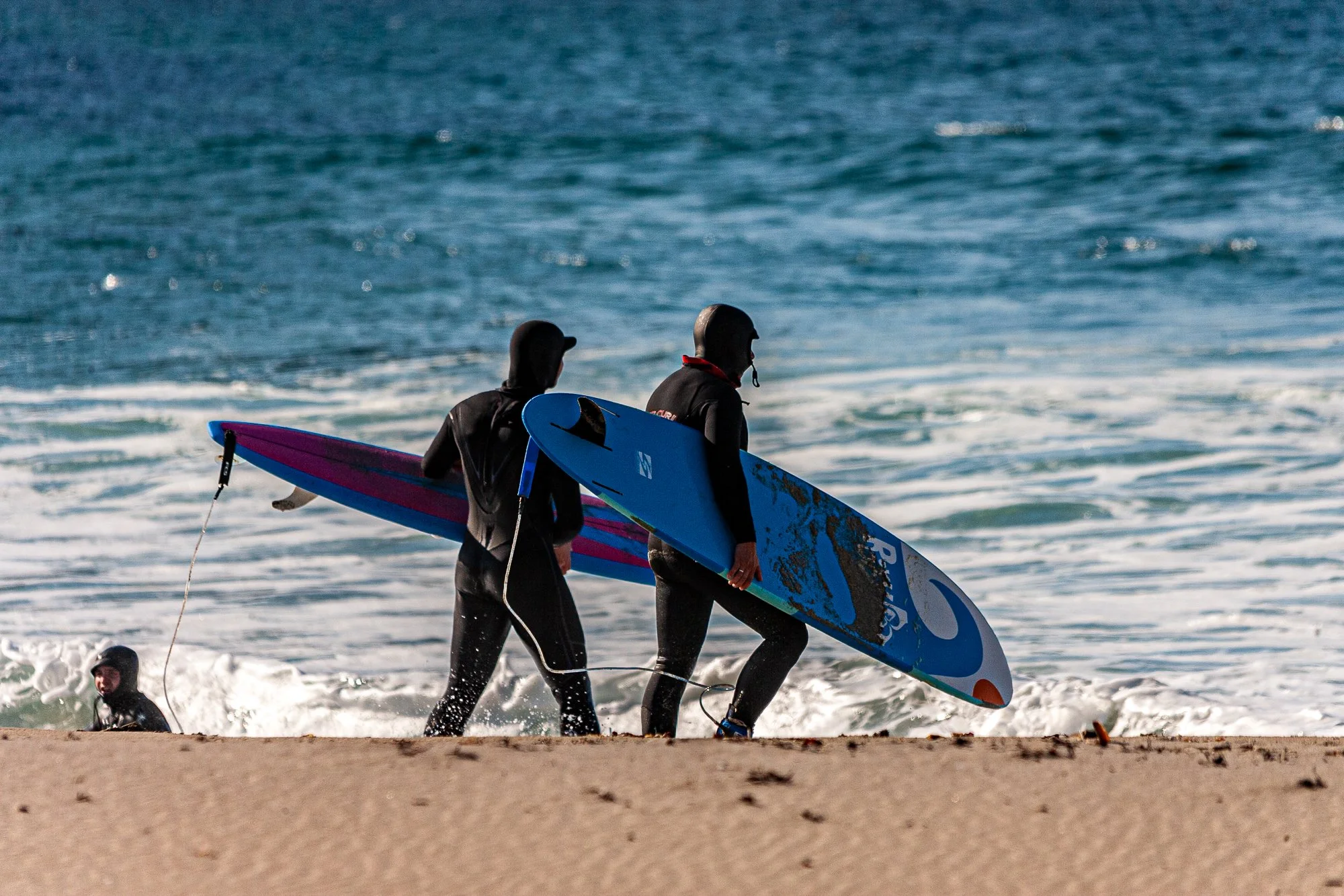 To personer i våtdrakter går ut av vannet på stranden med surfebrett.
