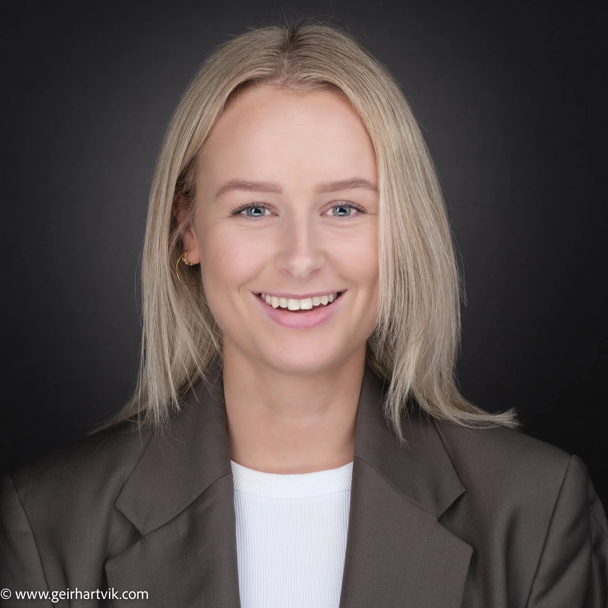 Profesjonelt portrett av en smilende kvinne med blond hår, iført grå blazer og hvit topp, mot en mørk bakgrunn.