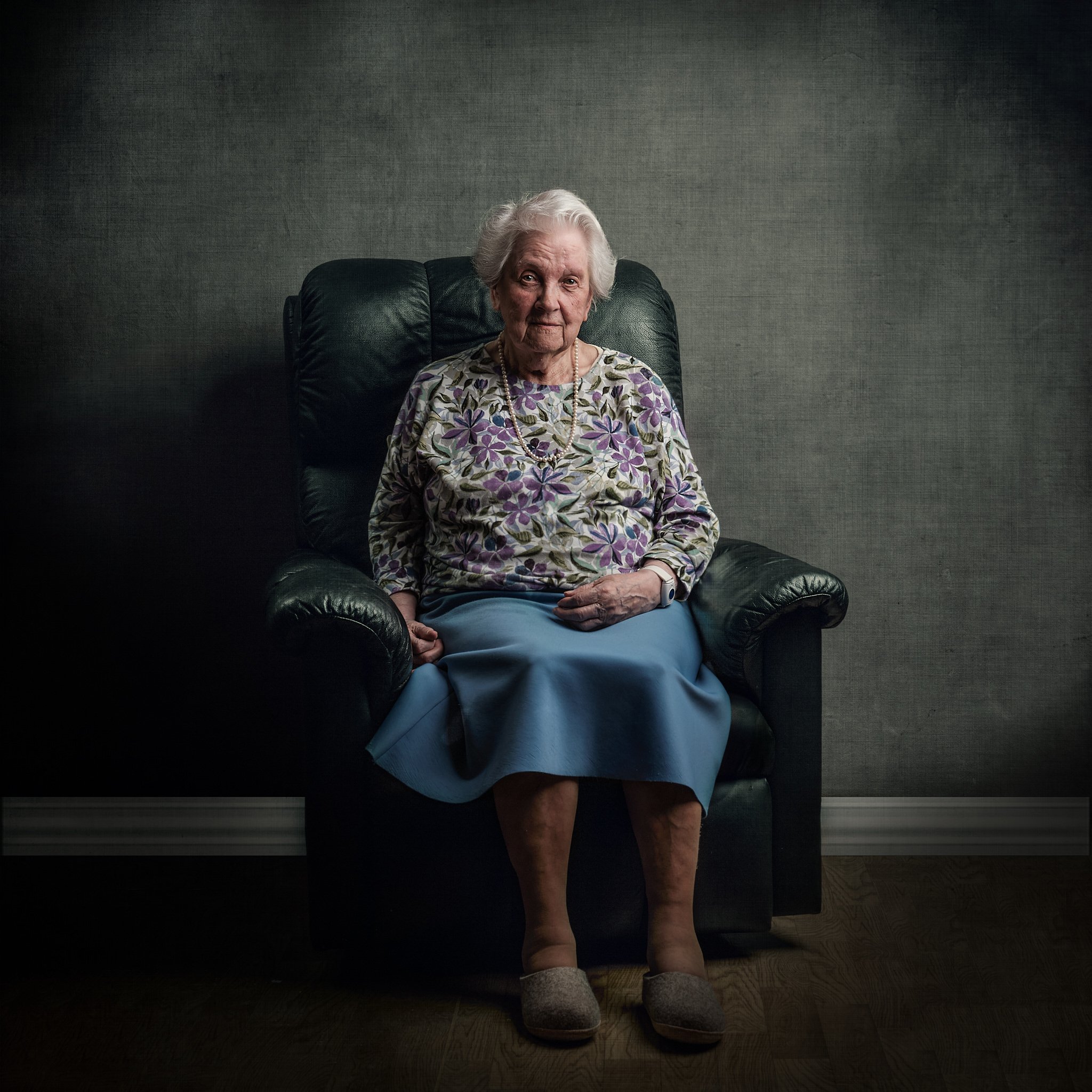 Eldre kvinne sitter i en svart lenestol mot en mørk grå vegg, iført blomstrete genser og blå skjørt, med smykker og tøfler.