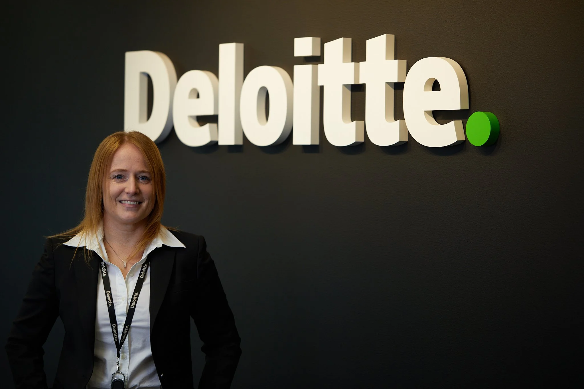 En kvinnelig person står foran en vegg med 'Deloitte'-logoen og et grønt punkt, smilende i profesjonell bekledning med svart jakke og hvit skjorte.