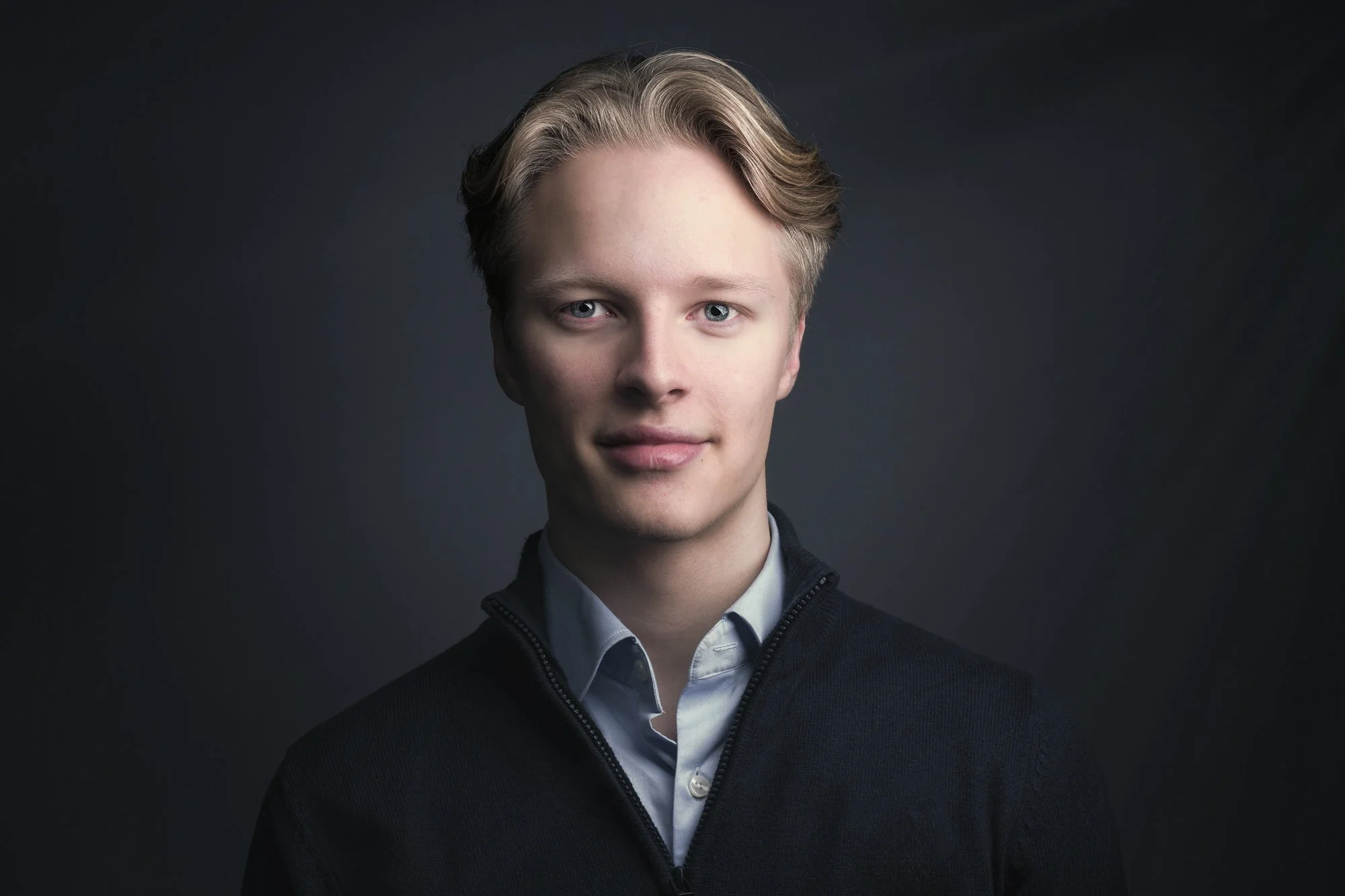 Ung mann med lyst hår og blå øyne, iført mørk genser, fotografert mot mørk bakgrunn.