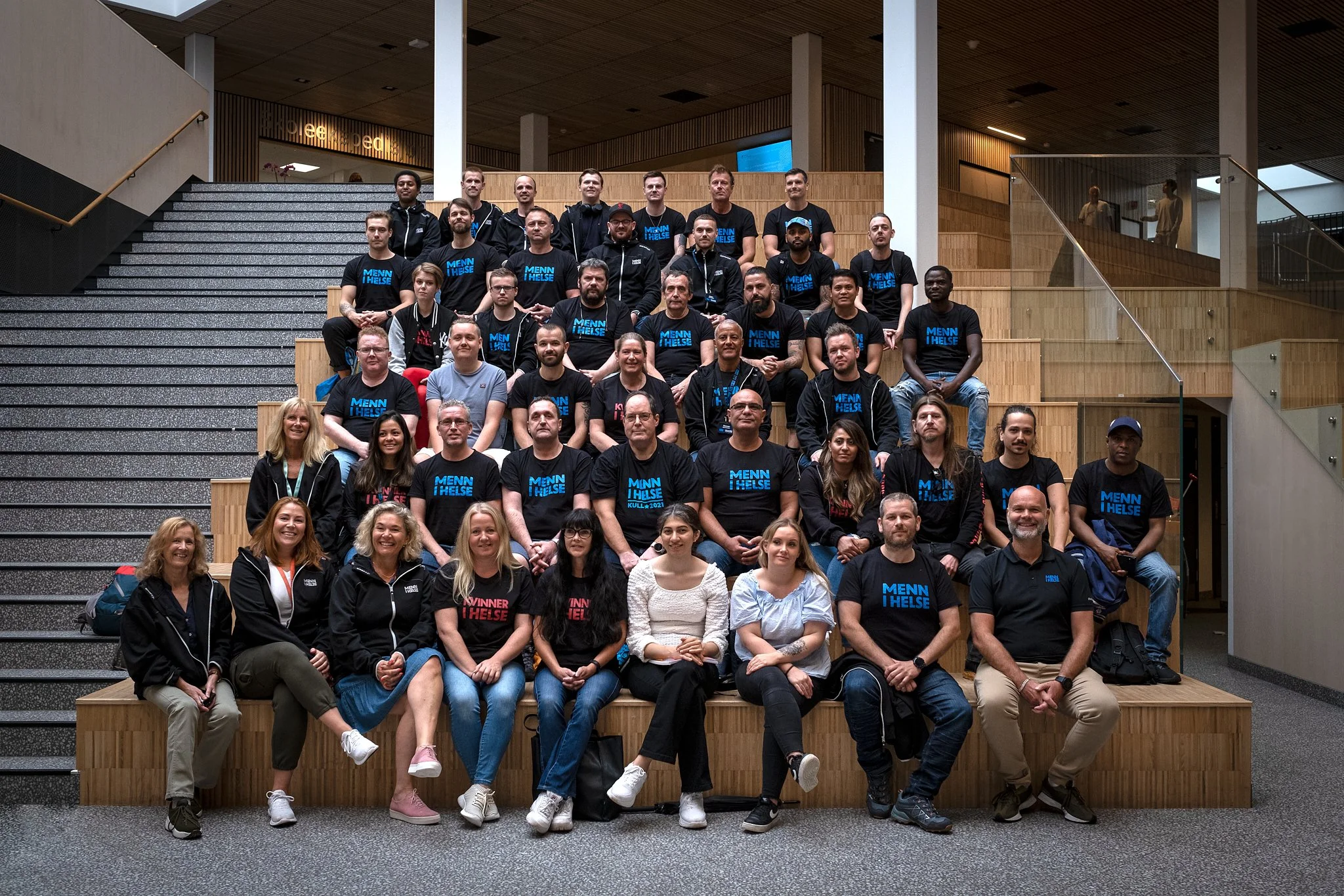 Gruppe av mennesker som sitter og står på trappetrinn i en bygning, mange av dem bærer svarte t-skjorter med teksten 'MENN I HELSE'.