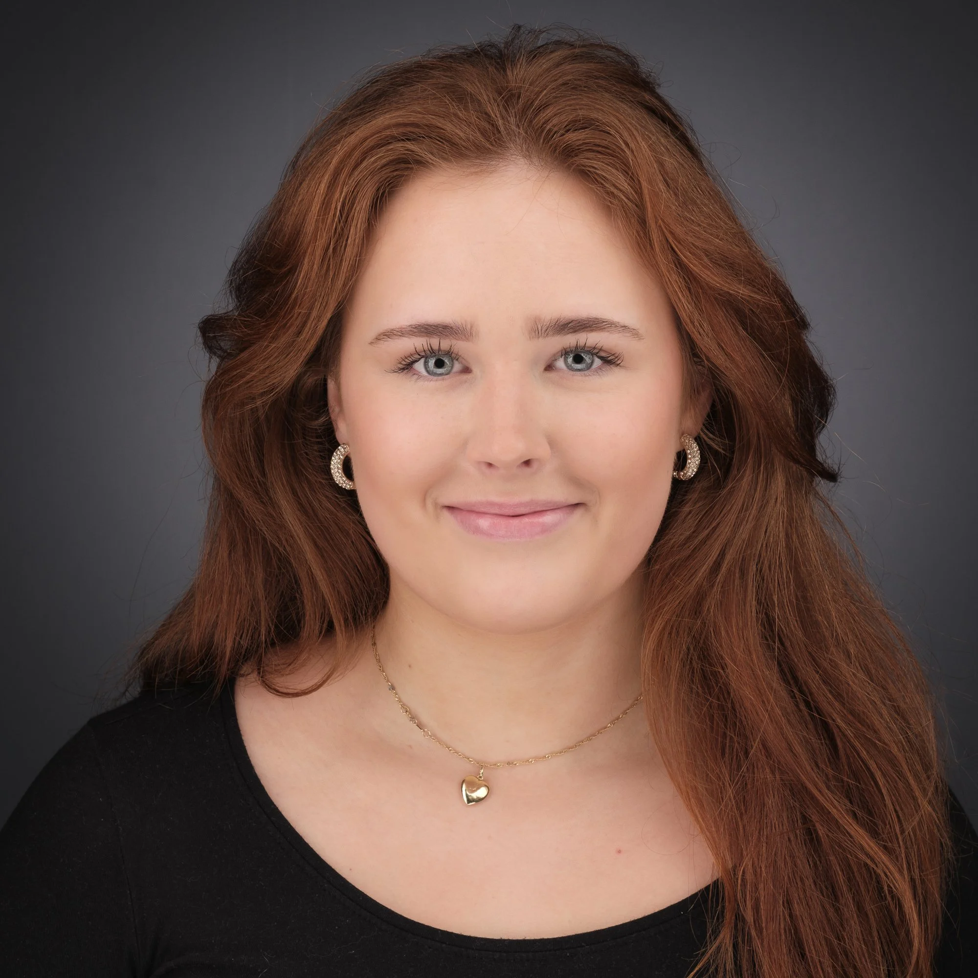 Portrett av en kvinne med rødt hår, smilende, iført svart topp og gullsmykker, mot en grå bakgrunn.