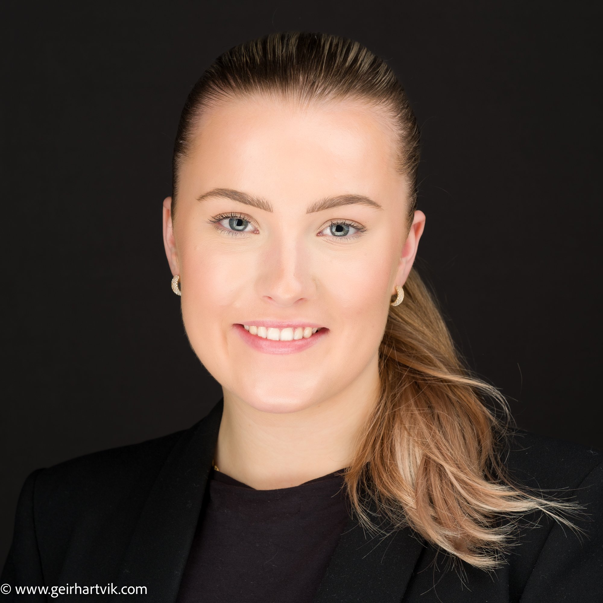 Portrett av en smilende kvinne med langt, lyst hår og blå øyne, iført svart blazer og small øredobber, mot en svart bakgrunn.