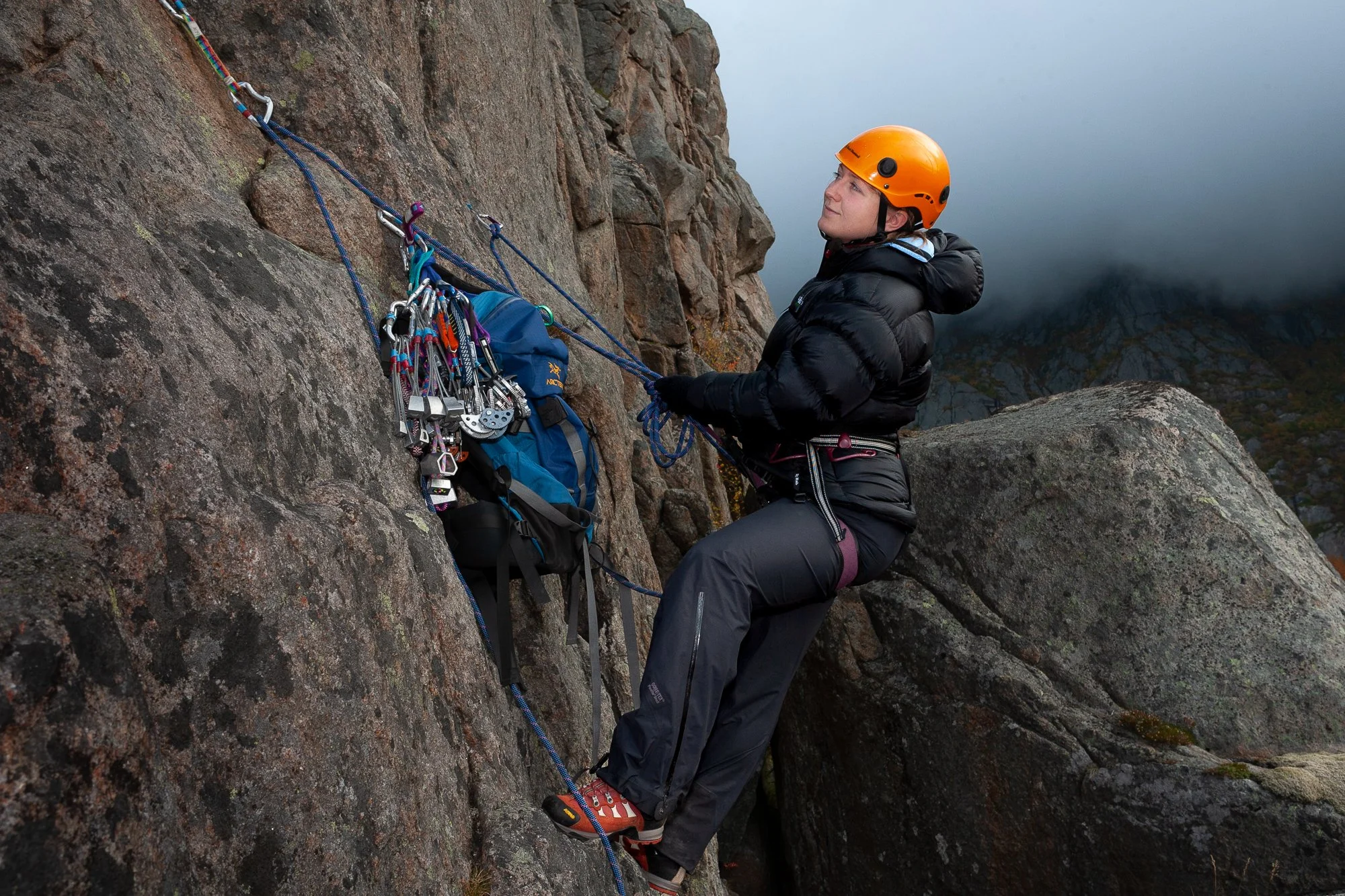 En kvinne med hjelm og fjellklatringsutstyr som står på en fjellvegg og ser opp.