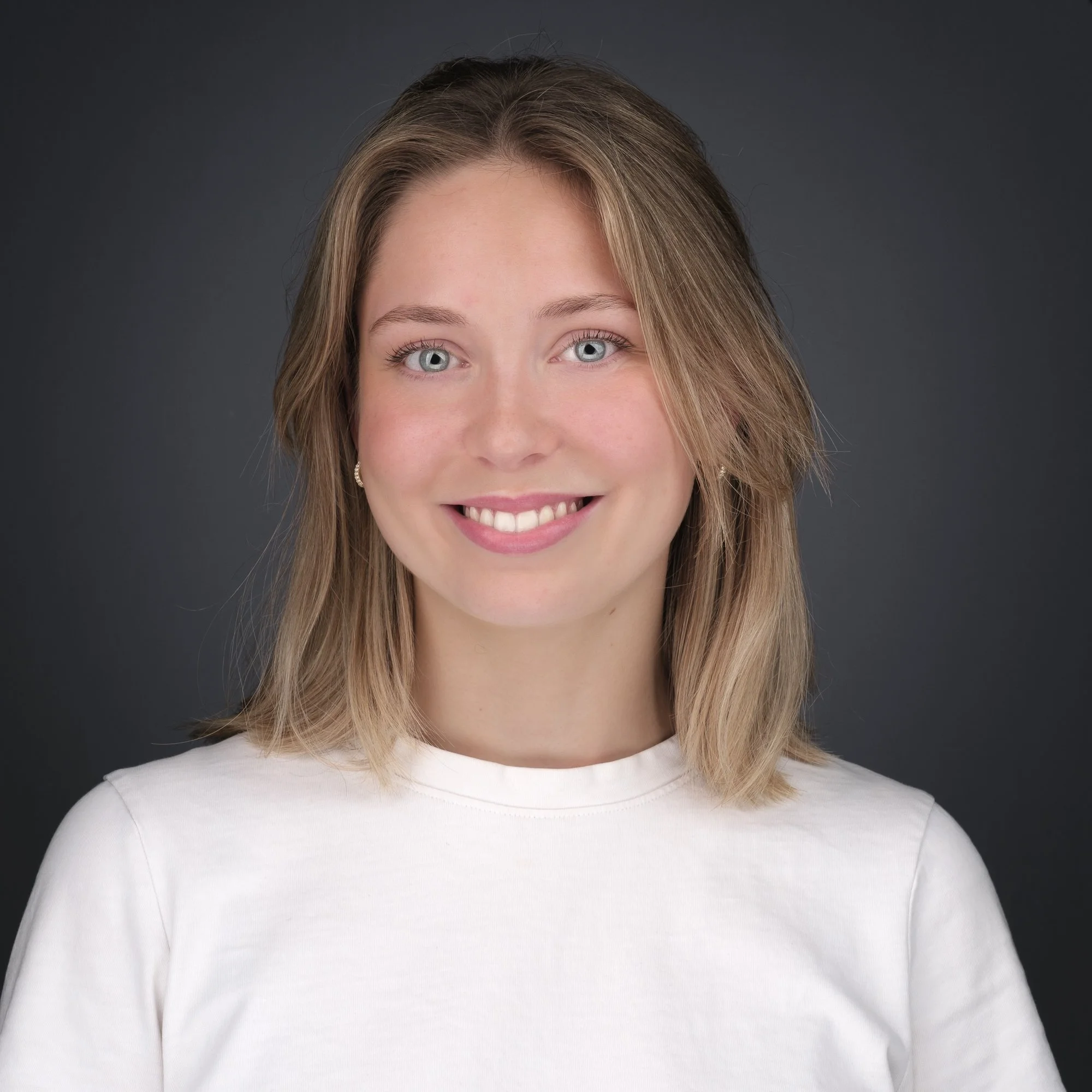 Portrett av en smilende ung kvinne med skulderlangt hår og hvit t-skjorte mot en mørk bakgrunn.