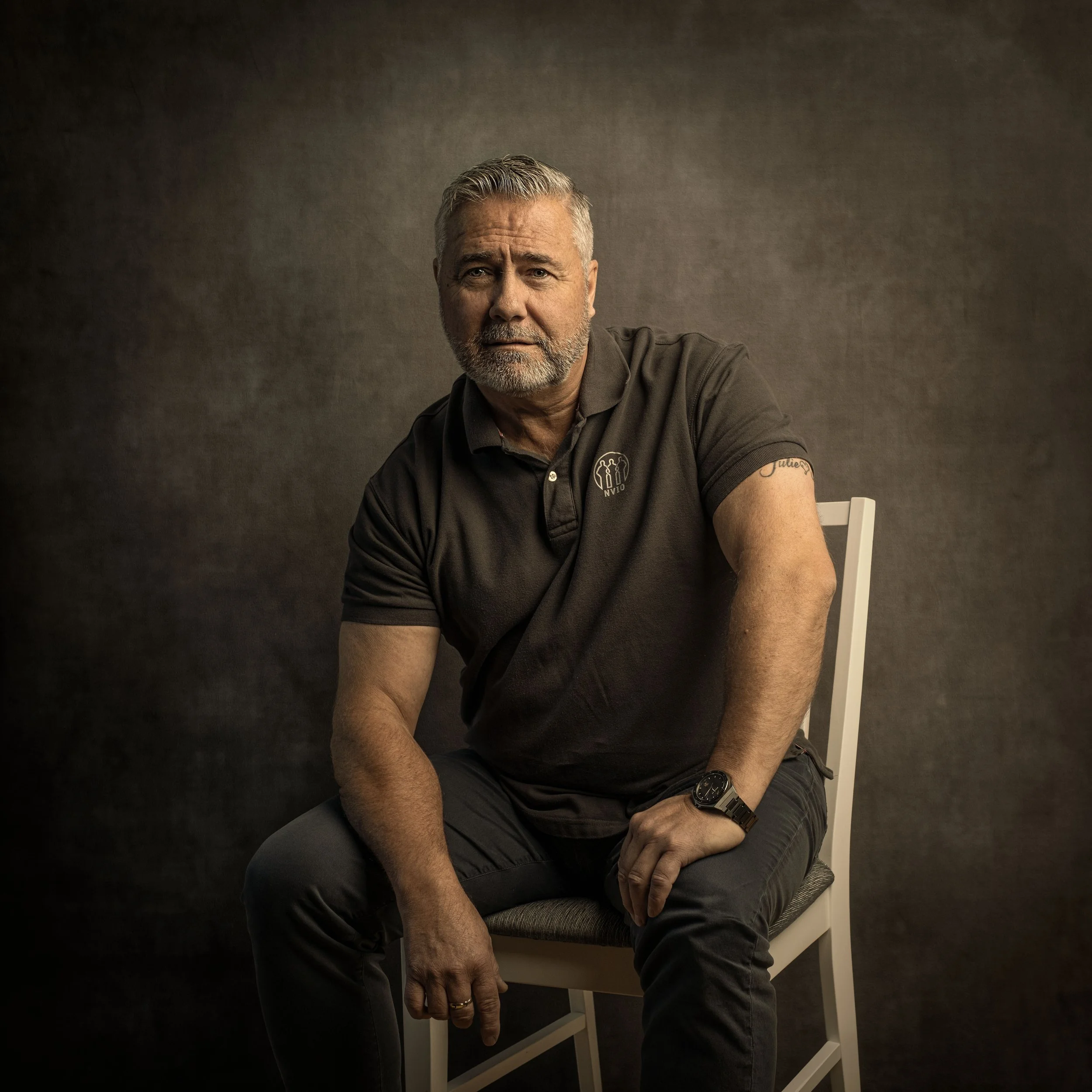 En eldre mann med grått hår og skjegg som sitter på en stol mot en mørk bakgrunn. Han har på seg en svart polo med et lite ikon på brystet, og har en svart klokke på venstre hånd.