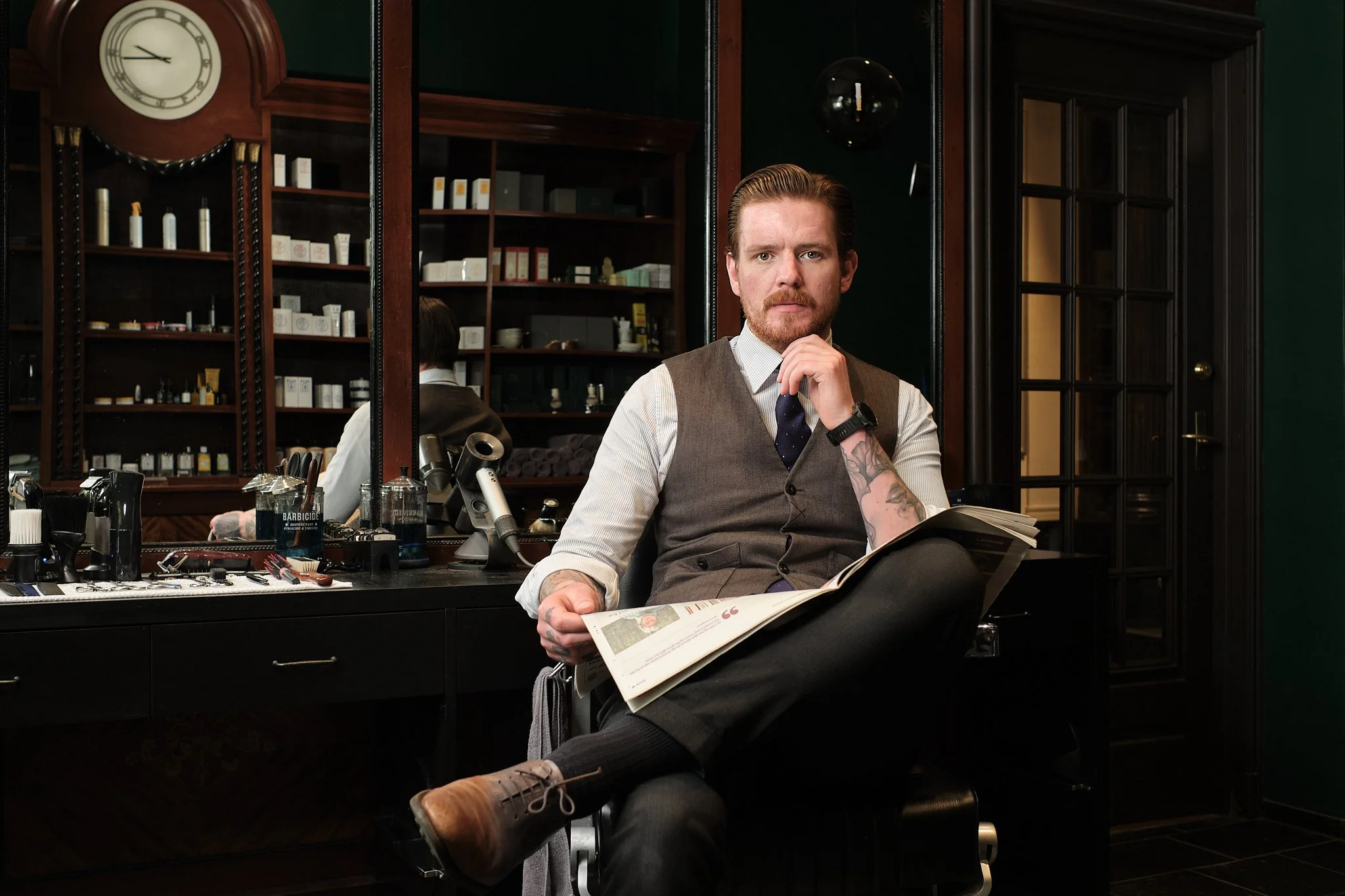 En mann med tatoveringer, iført vest og slips, sitter i en barbersalong. Han leser en avis og har en hånd under haken.