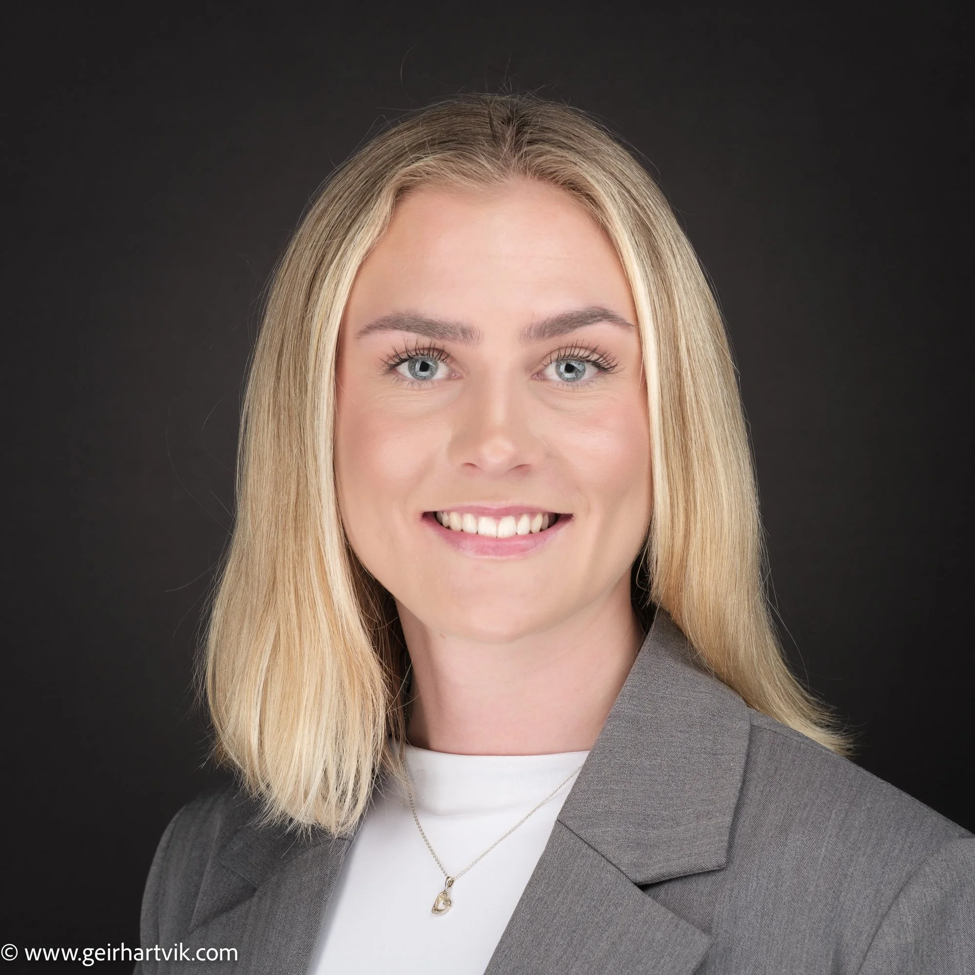 Nærbilde av en smilende ung kvinne med blond hår og blå øyne, iført dress og enkel smykker, mot en mørk bakgrunn.