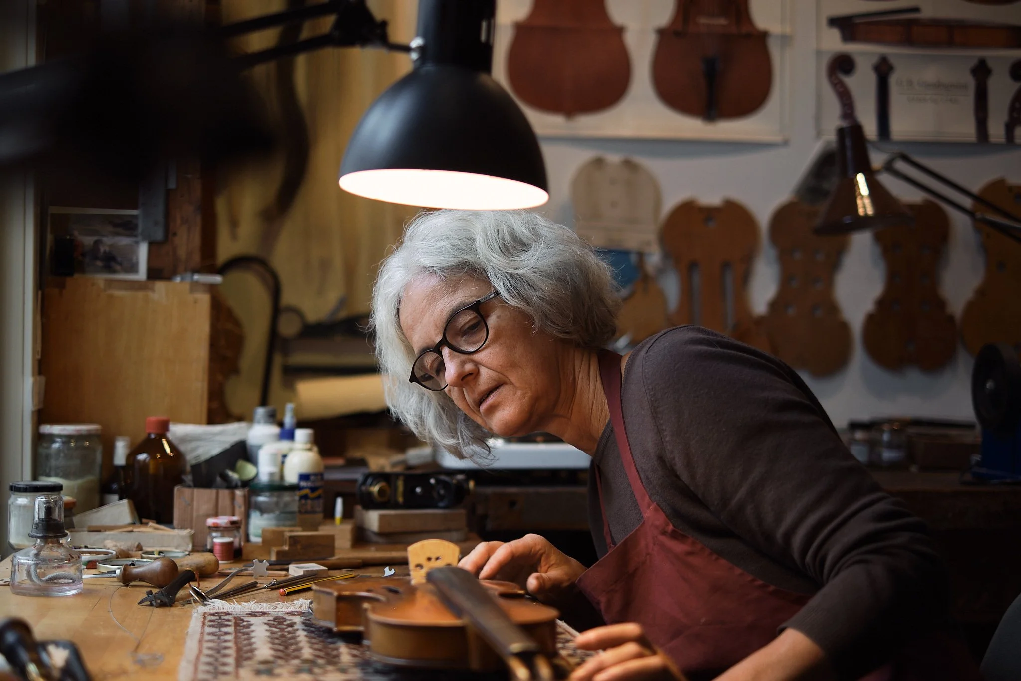En eldre kvinne med grått hår og briller jobber på et bord i et verksted, omgitt av ulike verktøy og materialer, med flere gitarer hengende på veggen bak henne.