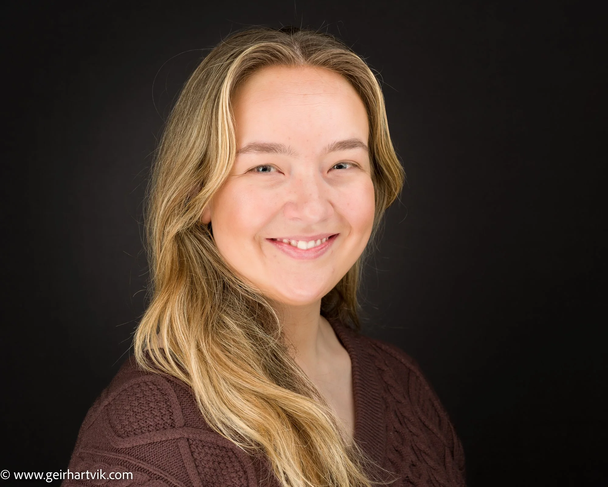 Portrett av en smilende kvinne med lang, krøllete, blond hår, iført en mørk rød genser, mot en svart bakgrunn.