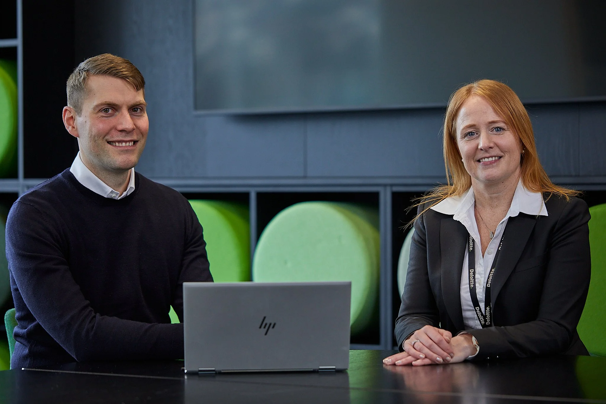 To personer som sitter ved et bord, en mann med lysebrunt hår og en kvinne med rødt hår, begge smilende, med en åpen laptop foran dem.