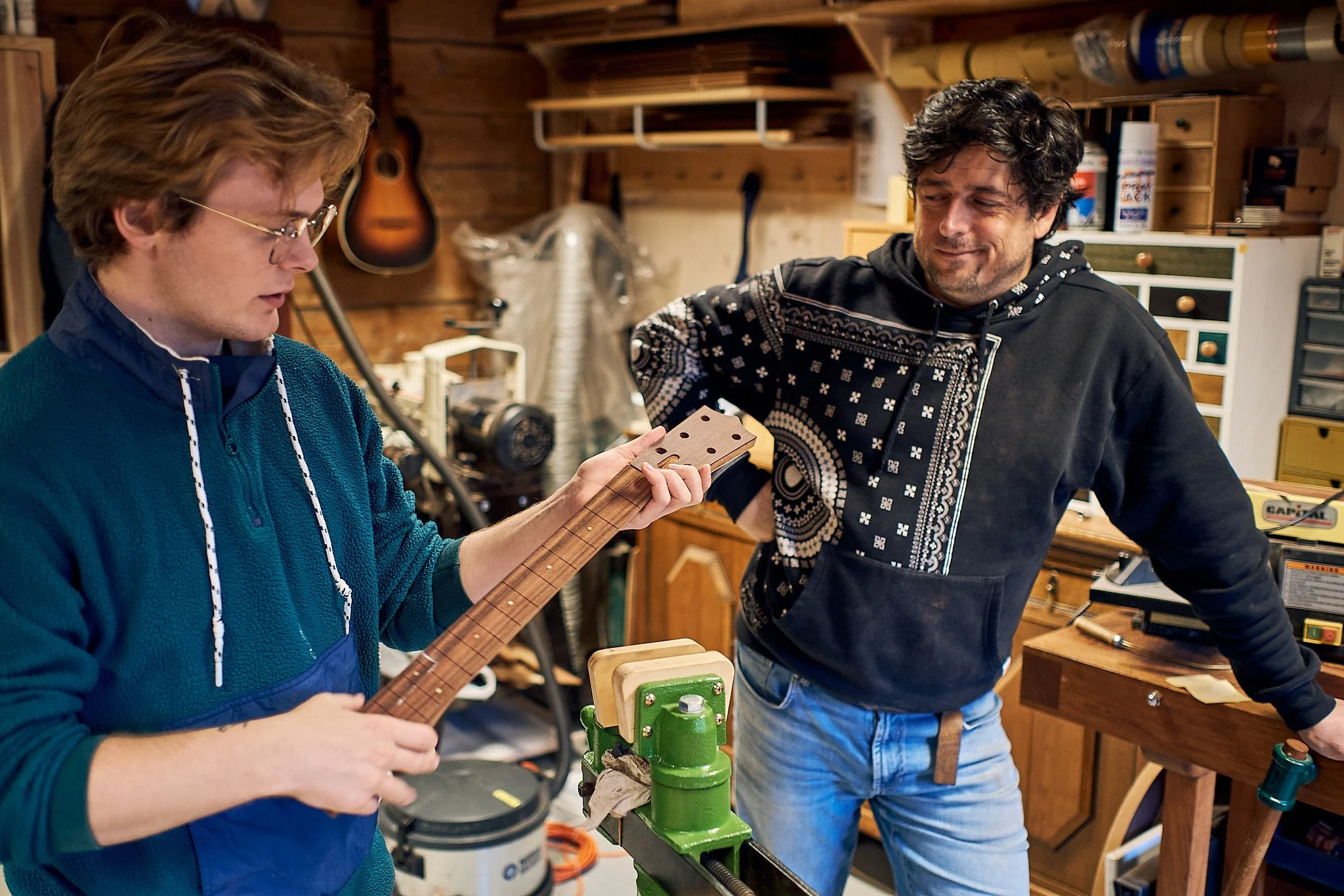 To menn i et trearbeverksmakersted. Den ene holder på en gitarhals av tre, mens den andre ser på og smiler.