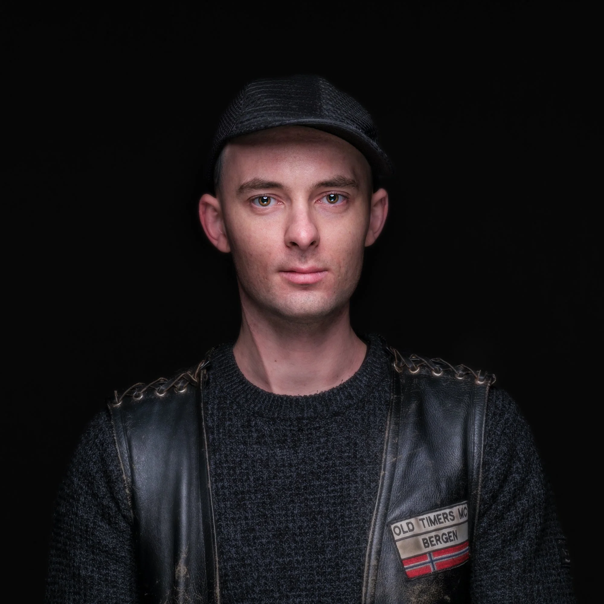 Portrett av en ung mann med lys hud, kort hår, blå øyne, iført svart lue, svart genser og skinnvest med ermestroper, mot en svart bakgrunn.