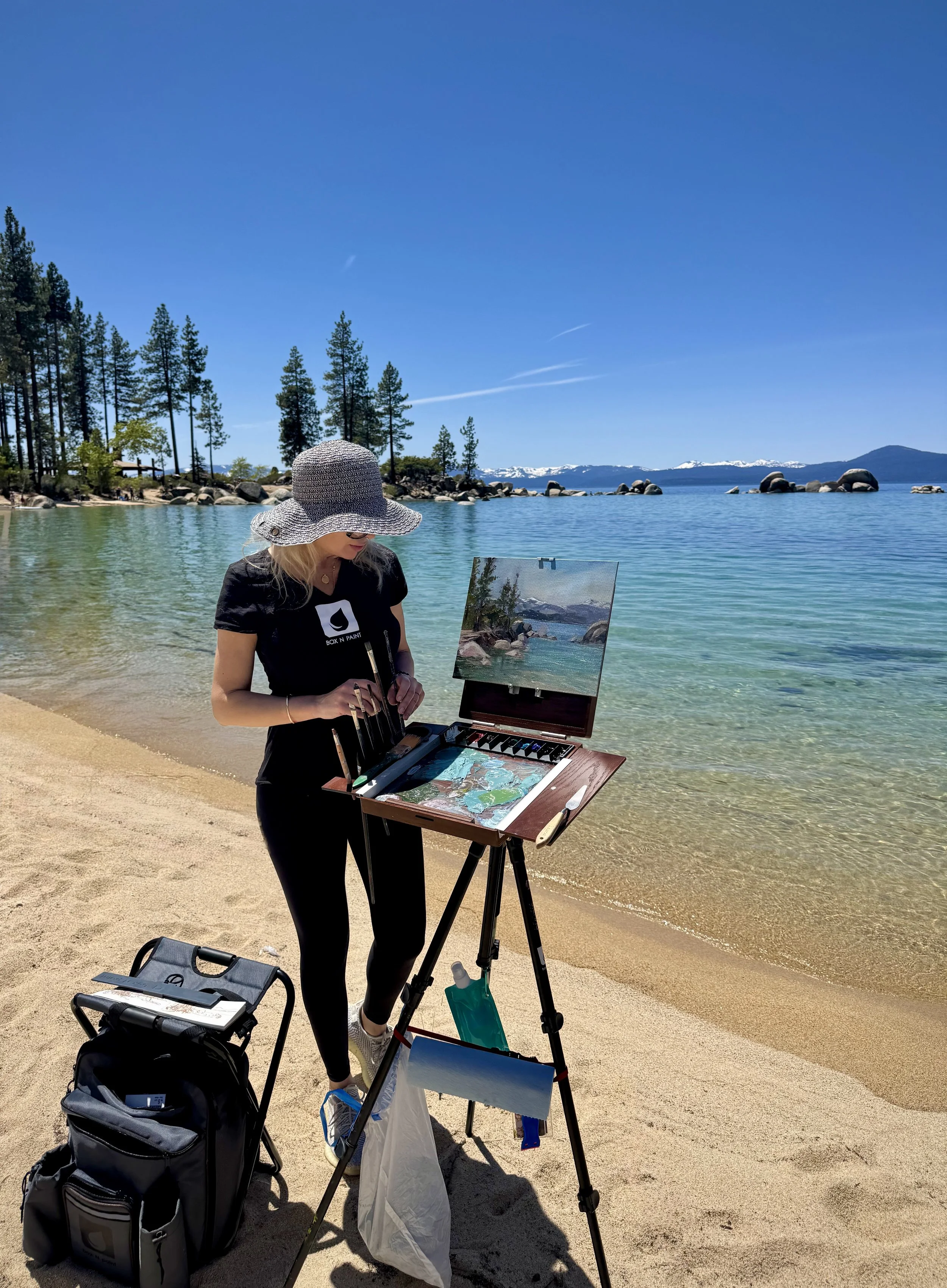 Woman painting landscape in an outdoor setting by a lake on a sunny day, with pine trees and snow-capped mountains in the background.
