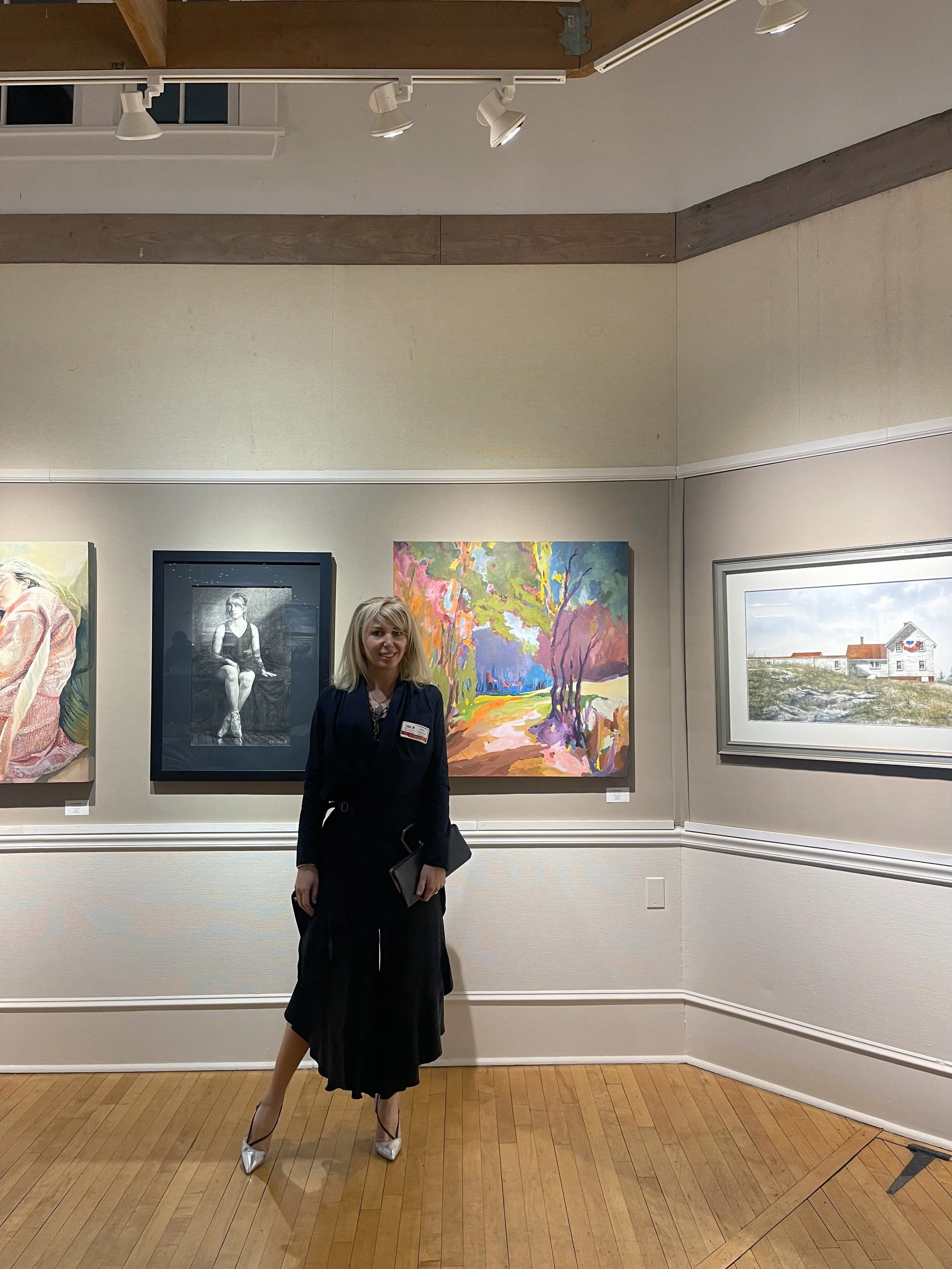 A woman with blond hair wearing a black dress and silver heels standing in an art gallery with various paintings on the wall behind her.