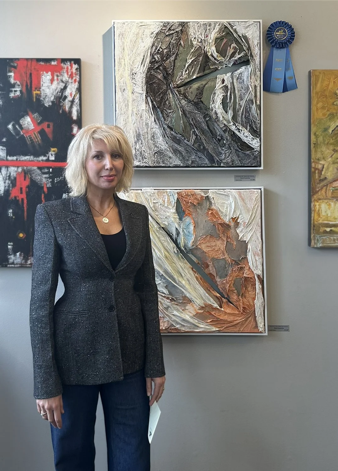 A woman with blonde hair wearing a black blazer and jeans standing in front of abstract artwork displayed on a gallery wall. There are multiple paintings, including black and red, gray with textured elements, and yellowish tones, with a blue ribbon a