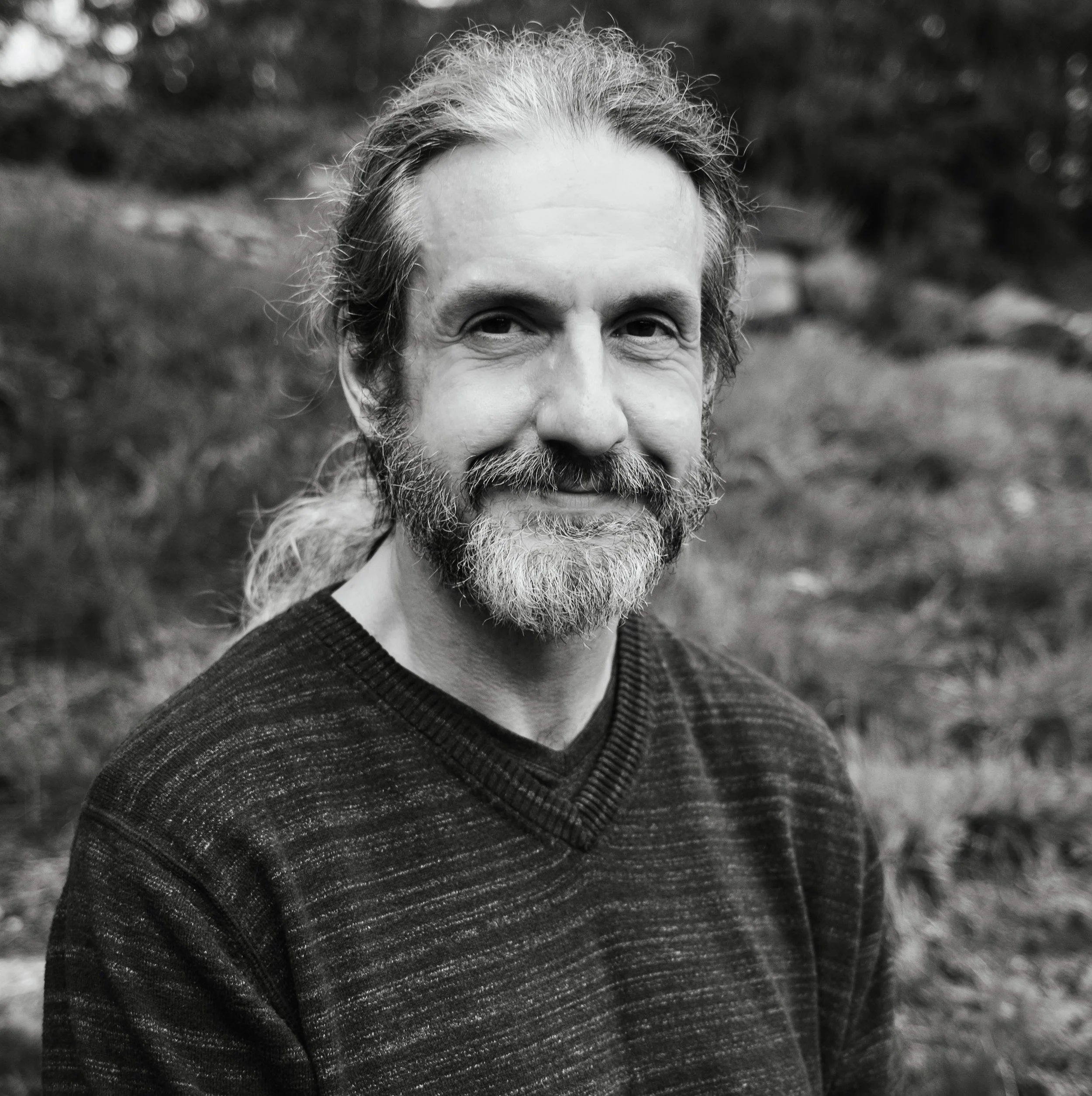 A black and white photo of a middle-aged man with a beard and long hair tied back, smiling outdoors with a blurred natural background.