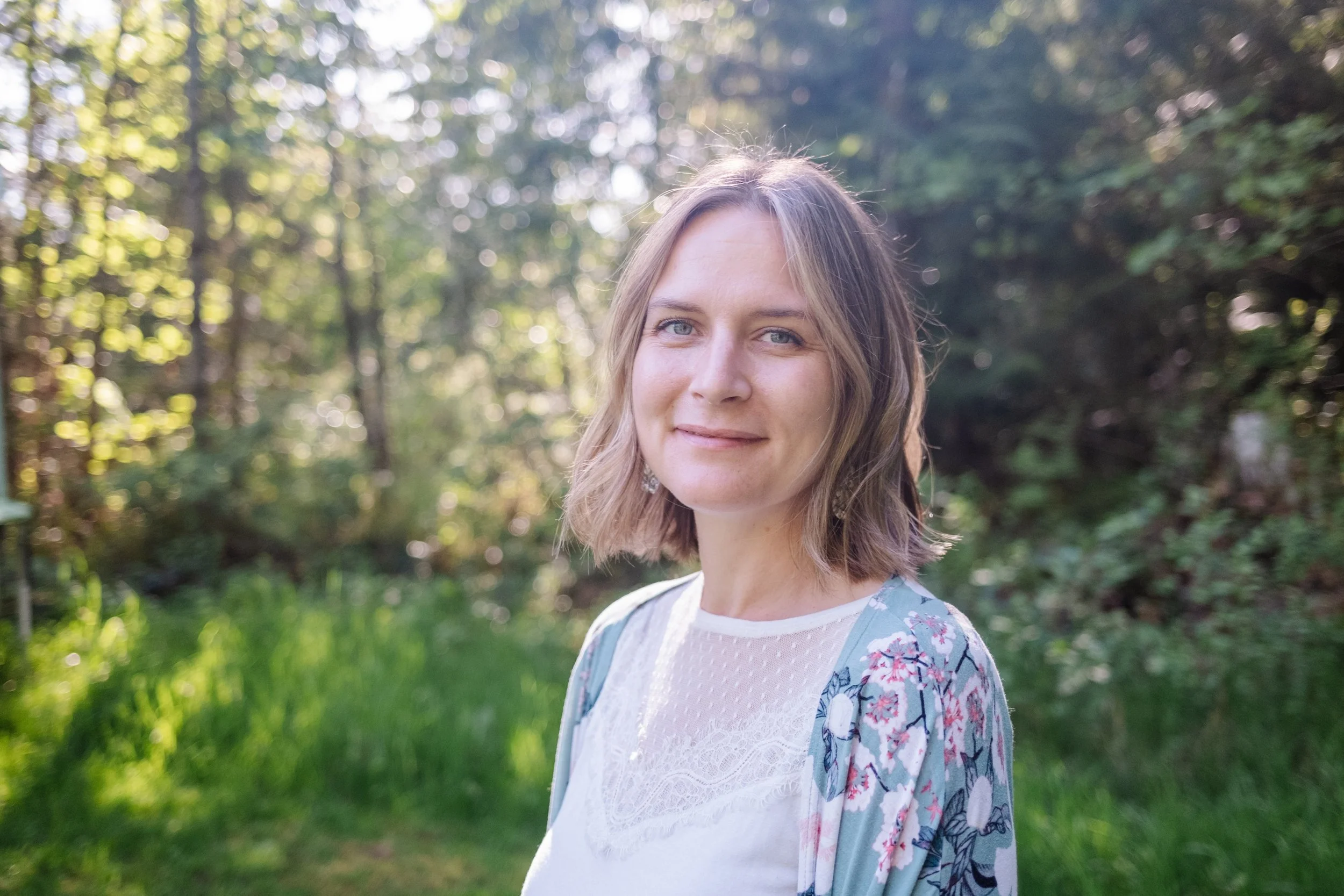 Smiling woman standing a natural blurry background with backlighting. Looks warm and approachable.