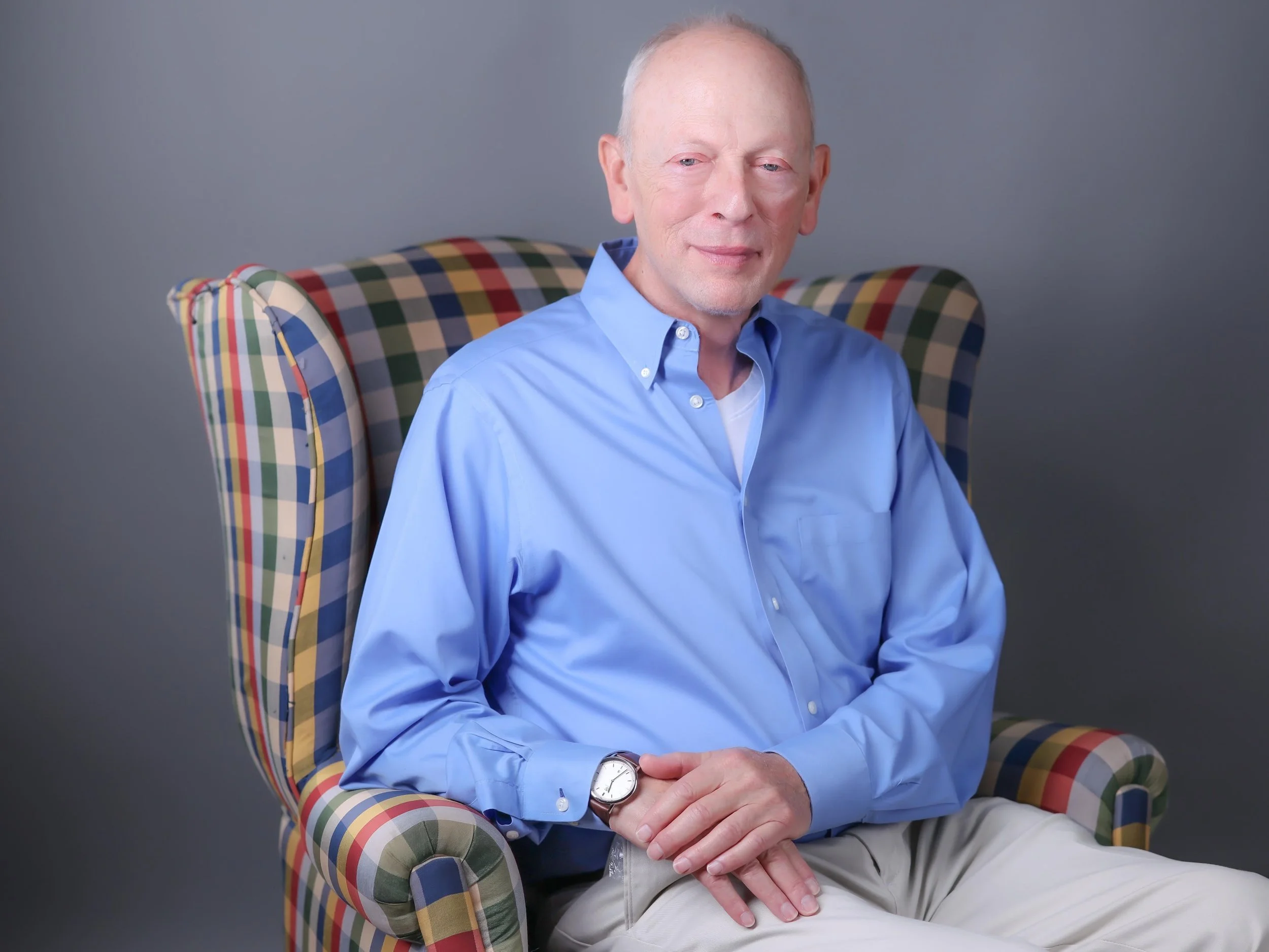 Photo of Dr. Harris wearing a blue button down shirt and sitting on a plaid chair