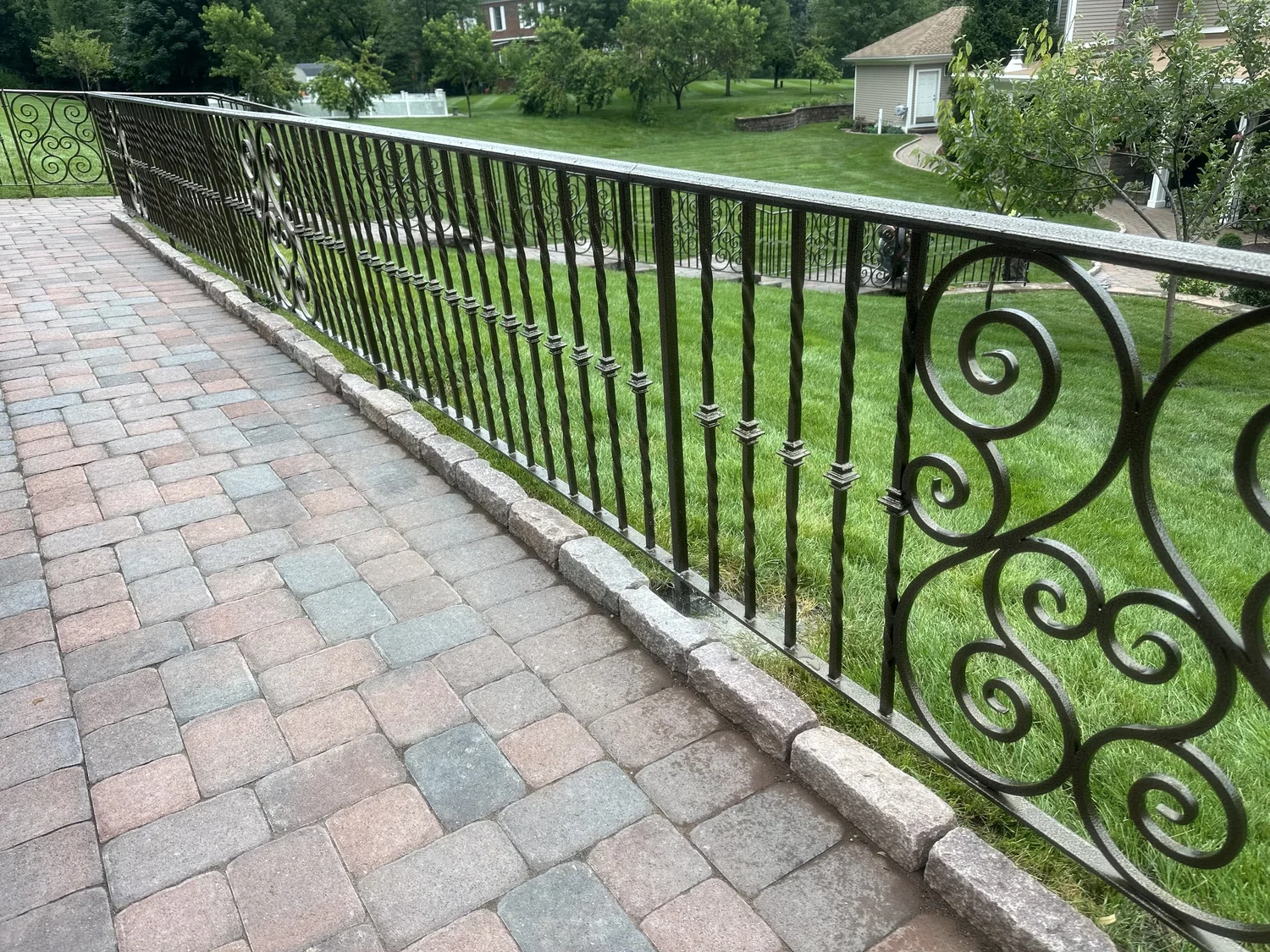 Ornamental scrollwork iron railing running along a brick paver walkway beside a lawn