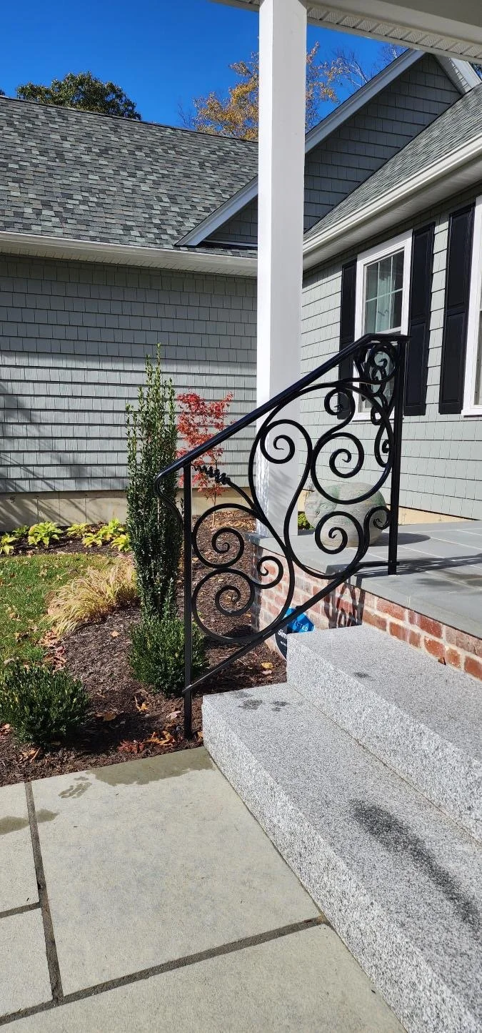 Residential house exterior showing a front porch with grey granite steps, a decorative black wrought iron railing, and a garden bed with small plants. Part of the house with grey siding, black shutters, and a window is visible, along with a part of the roof and clear blue sky in the background.
