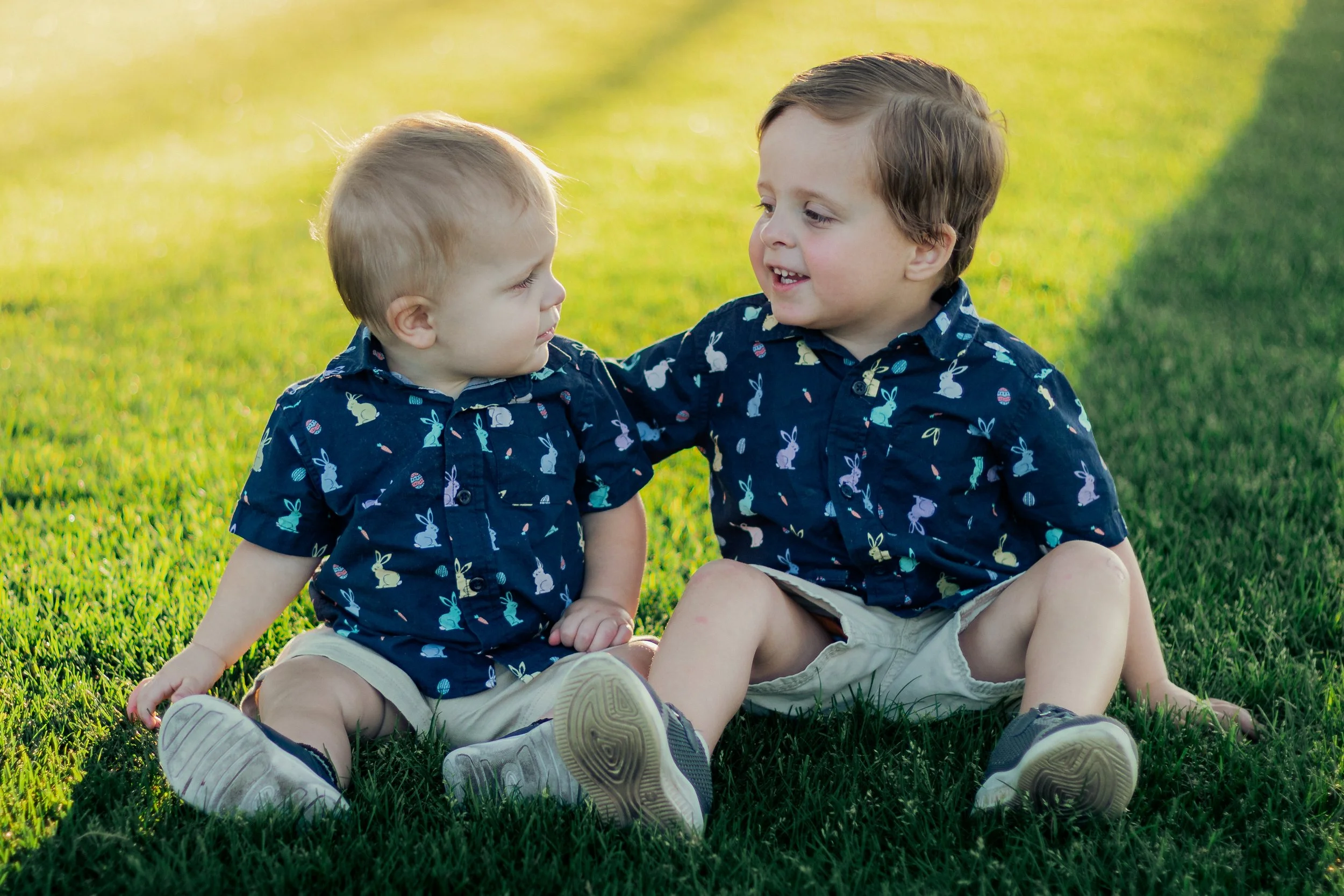 Two young boys sitting on grass in a park, wearing matching dark blue shirts with bunny patterns, and smiling at each other during the daytime.