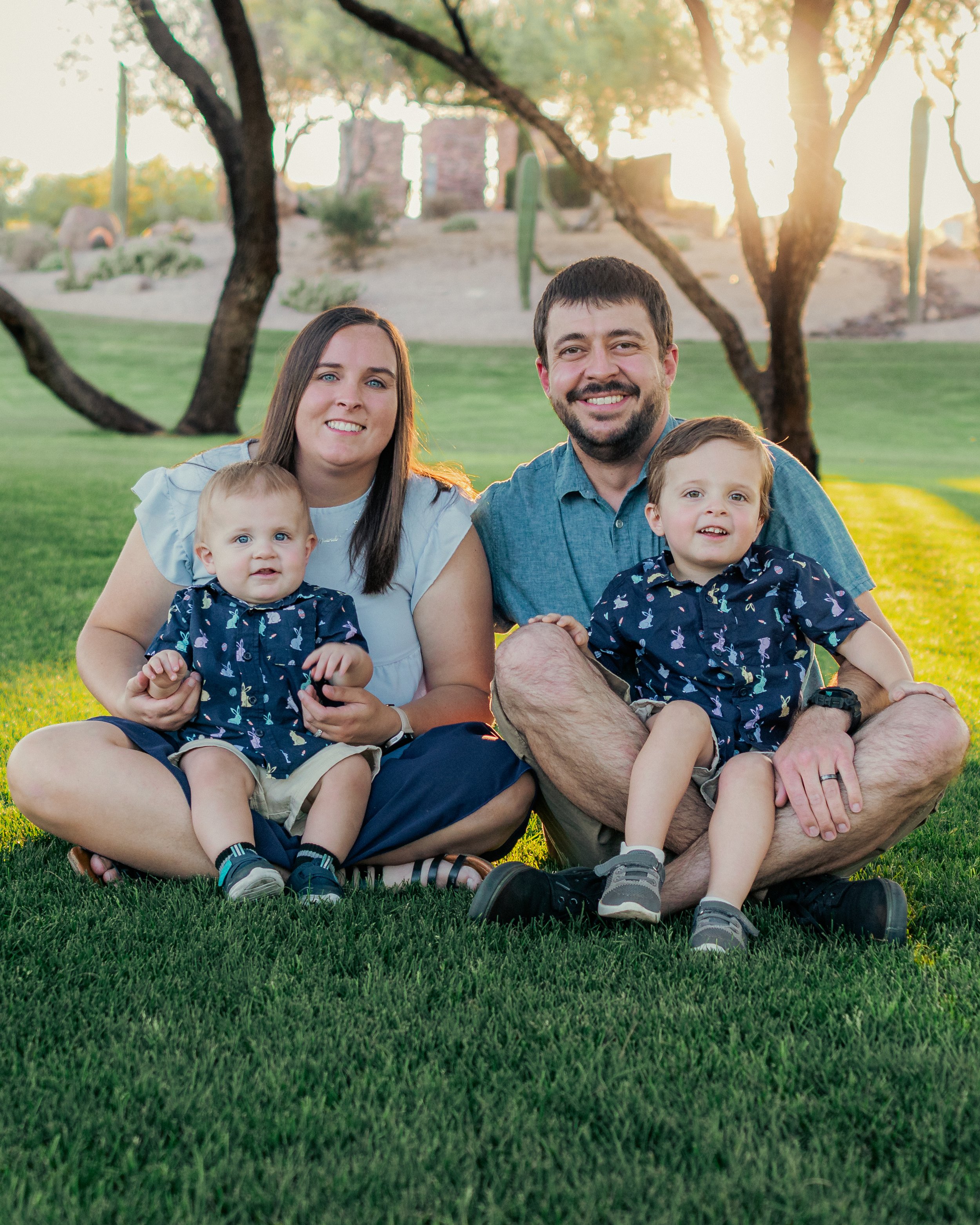 A family of four sitting on grass in a park during sunset, smiling at the camera. The family includes a woman, a man, and two young boys, all dressed casually, with trees and a gently sloping hillside in the background.