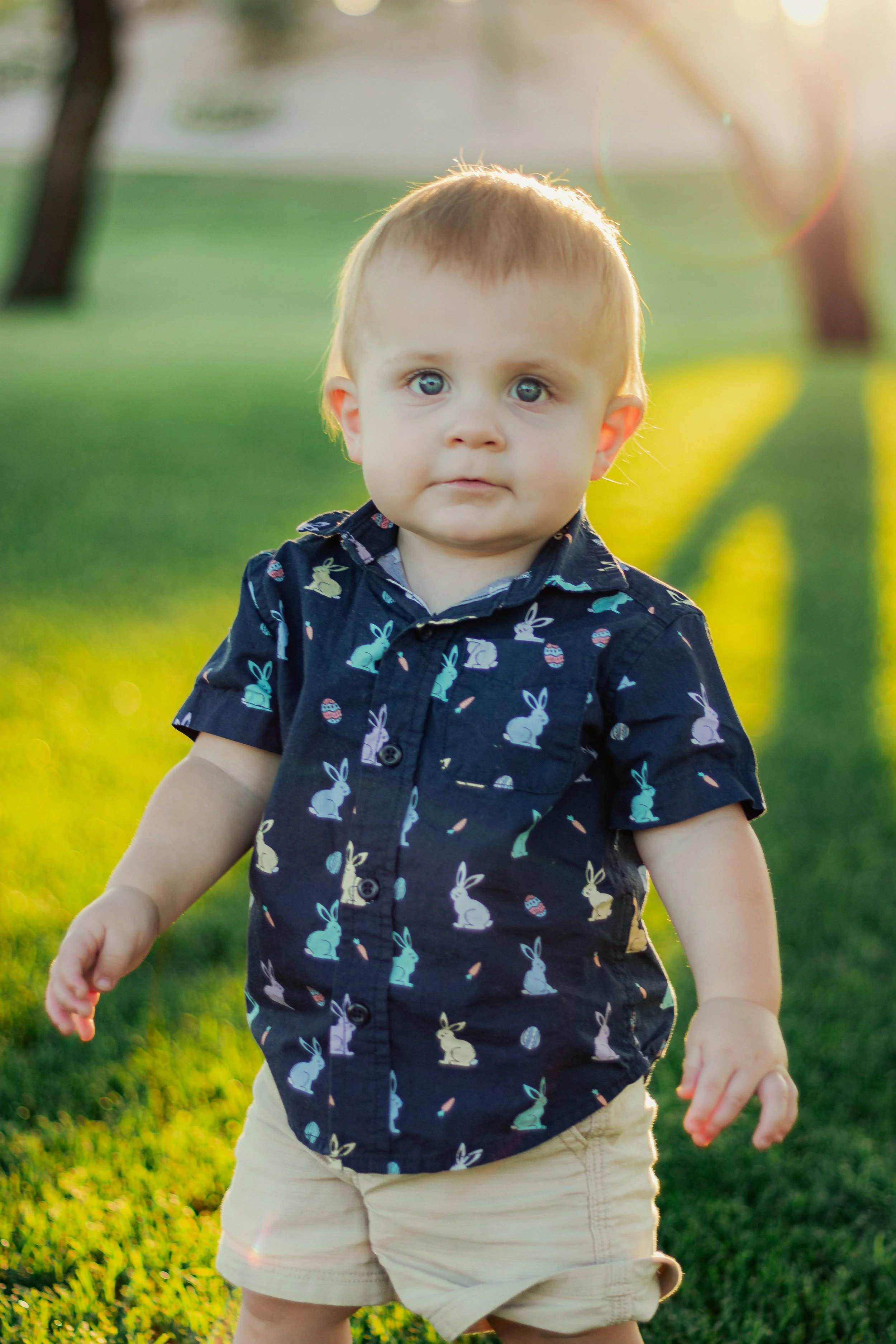 A young boy with light brown hair and blue eyes standing outdoors on a grassy field during sunset, wearing a navy blue shirt with pastel-colored bunny and egg patterns and beige shorts.
