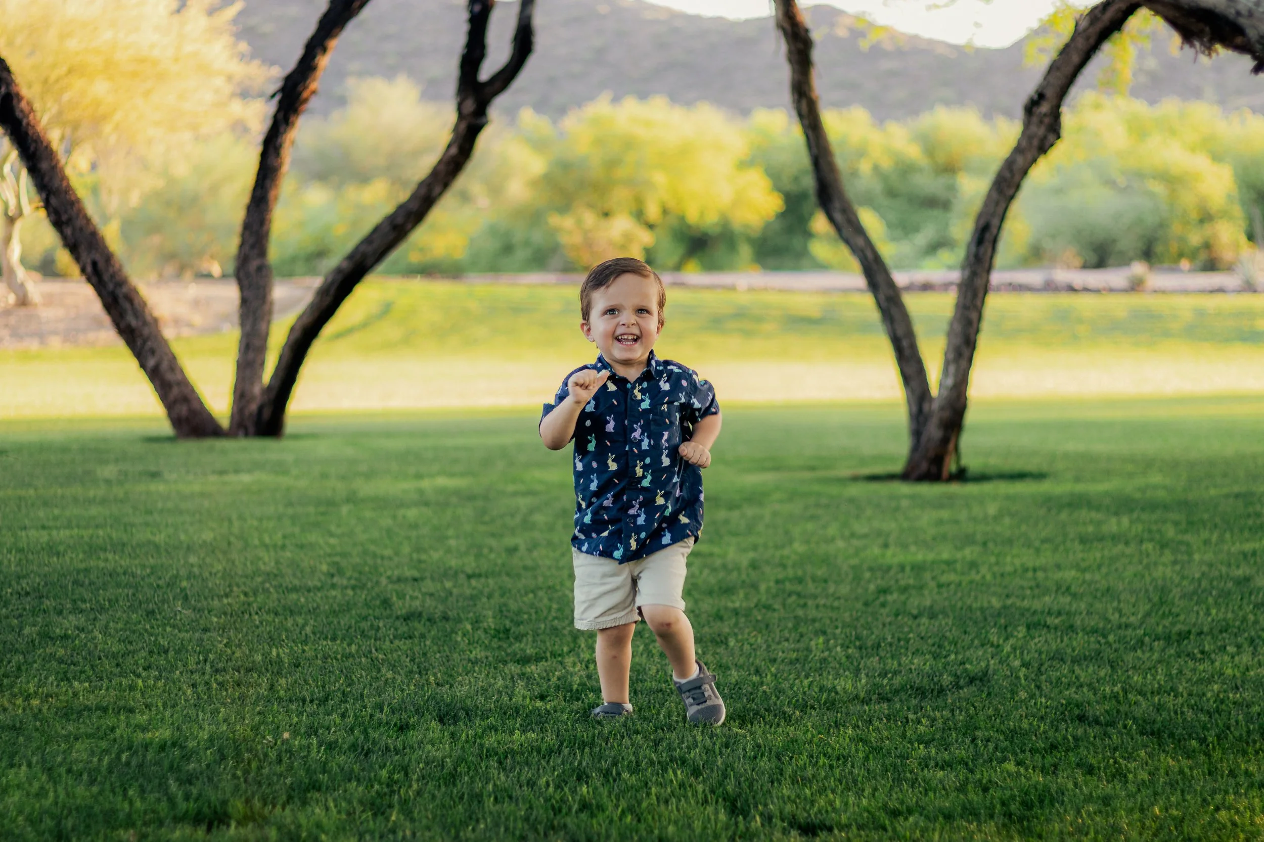 A young boy running on a lush green lawn with a big smile, wearing a navy blue shirt with colorful patterns and beige shorts, in a park with trees and a body of water in the background during daytime.