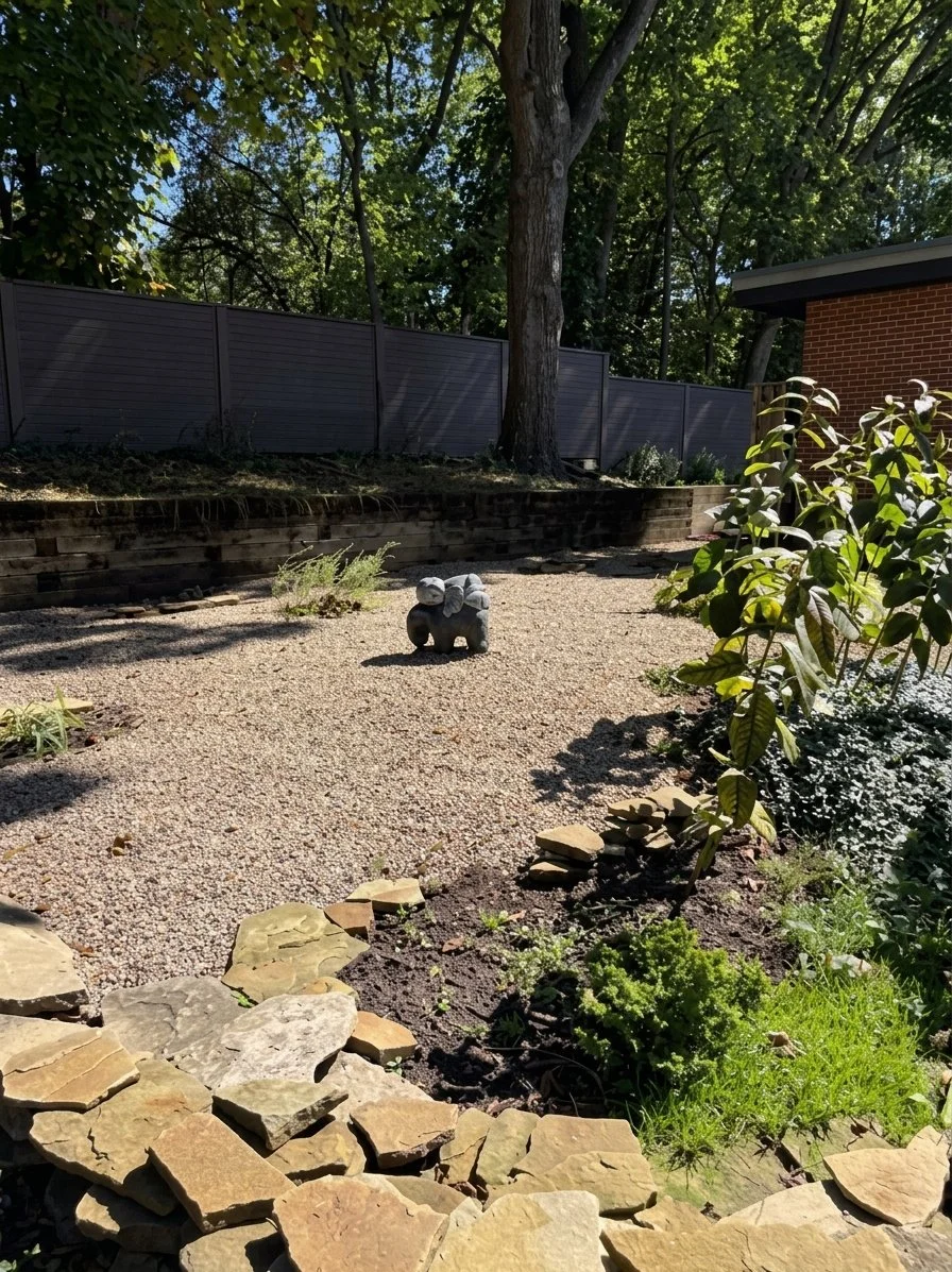 A backyard garden with a gravel pathway, a small stone statue of an animal, surrounding plants, a tree, a brick house, and a gray fence.