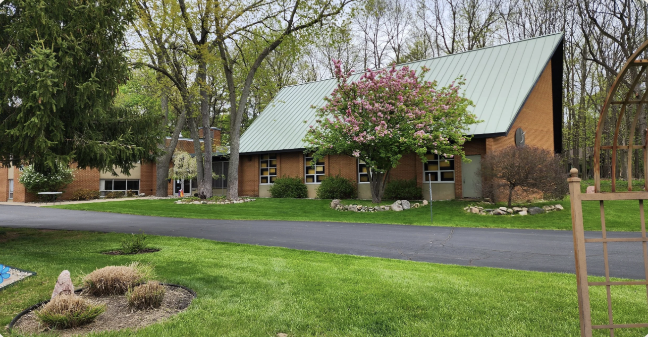 A brick church with a green metal roof, trees, and a grassy lawn with flower beds.