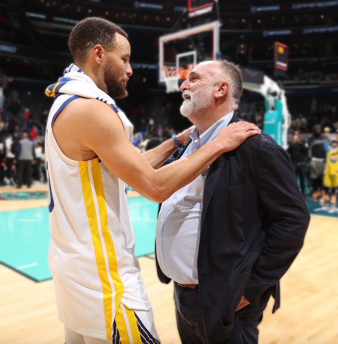 Stephen Curry and José Andrés share a moment on the basketball court, with the player wearing a white and yellow jersey and the man in a black jacket, surrounded by a crowd in the background.