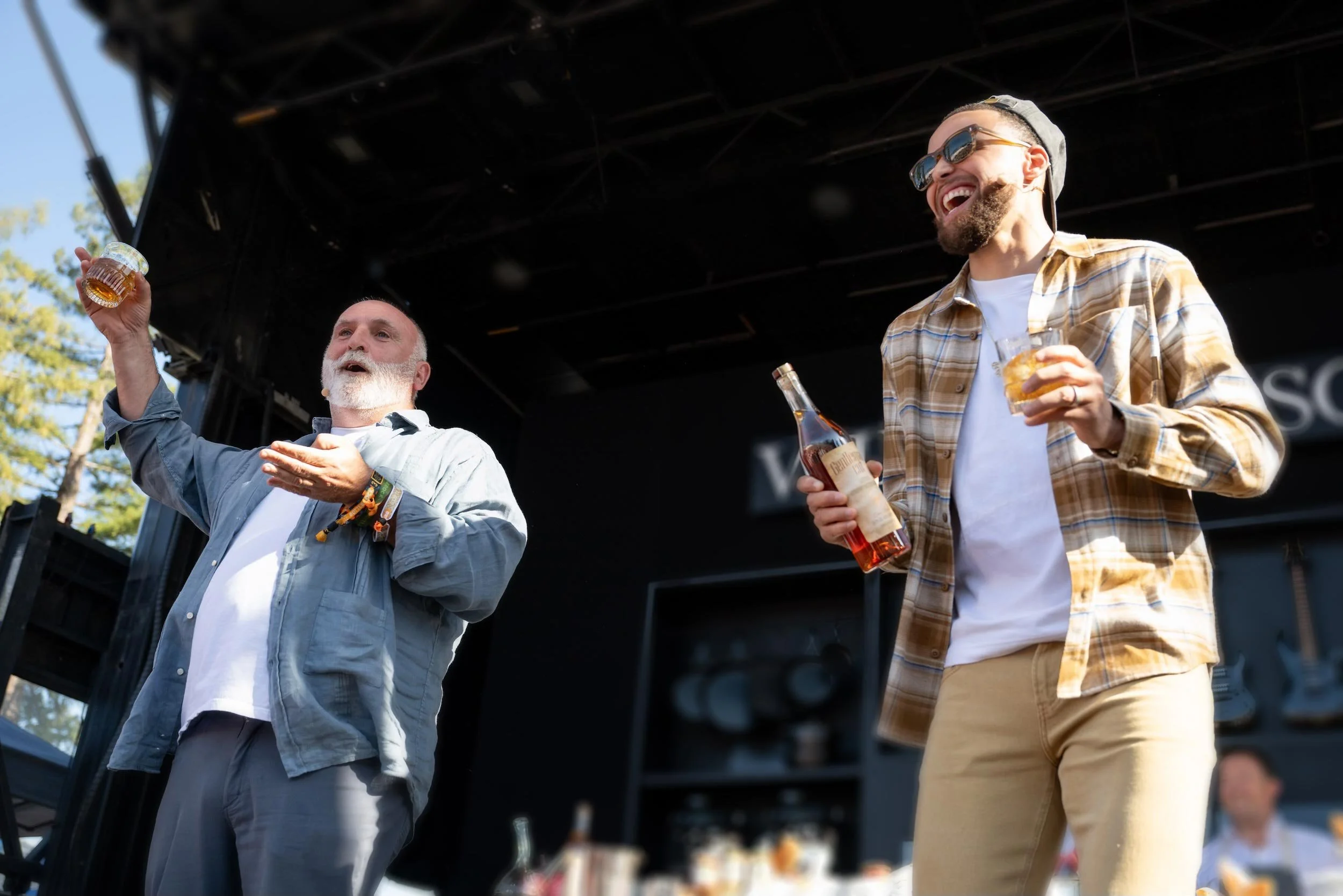 José Andrés with Stephen Curry on a stage, holding a bottle of Gentleman's Cut bourbon, both smiling and enjoying themselves at an outdoor event.