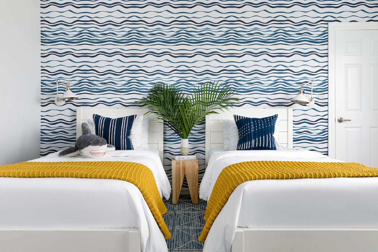 Ocean themed kids bedroom with blue and white wave wallpaper, white wooden bed frames, white comforters, blue throw pillows, and yellow throw blankets