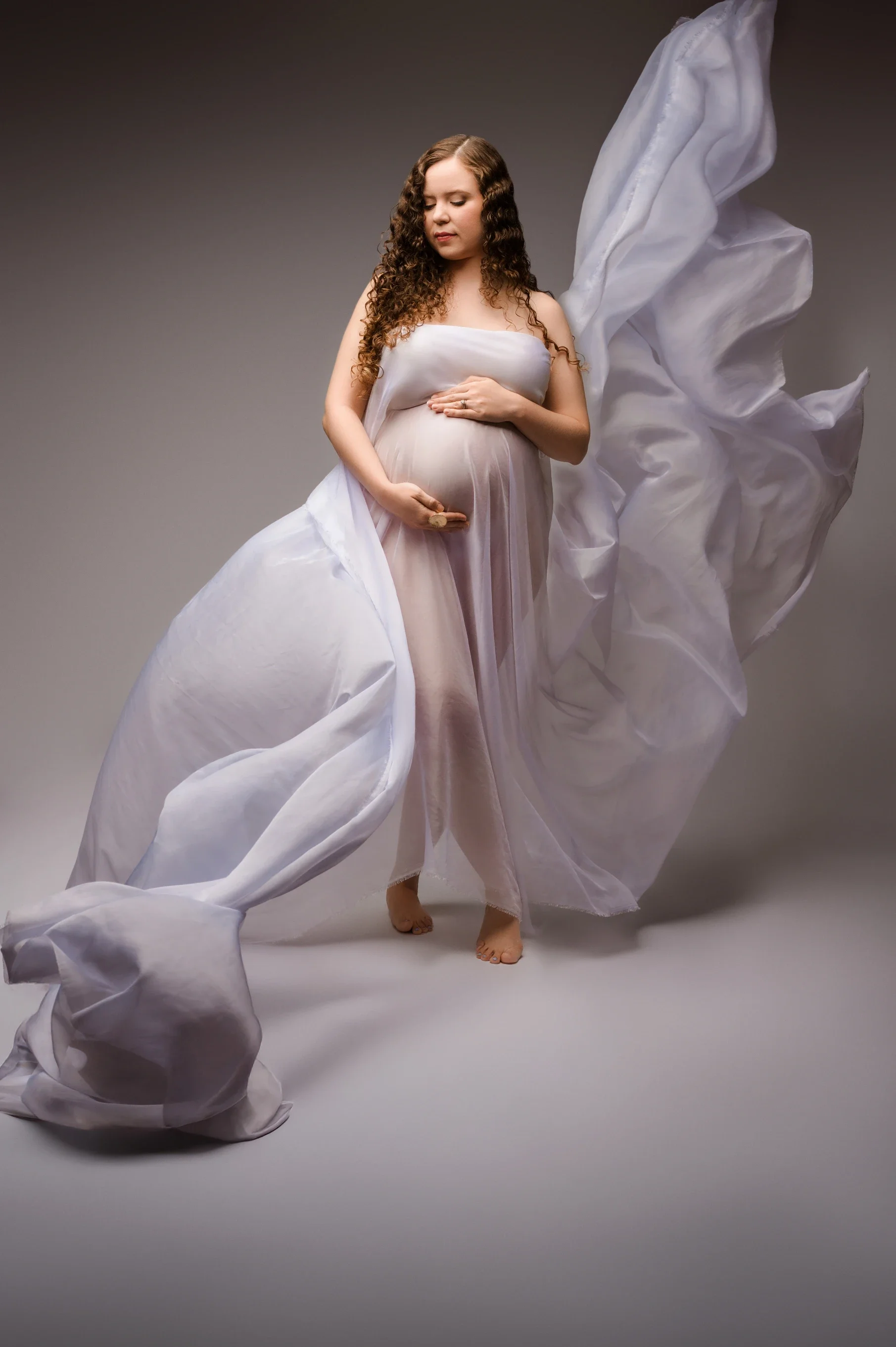 woman with curly hair holding her pregnant belly with flowing white fabric around her