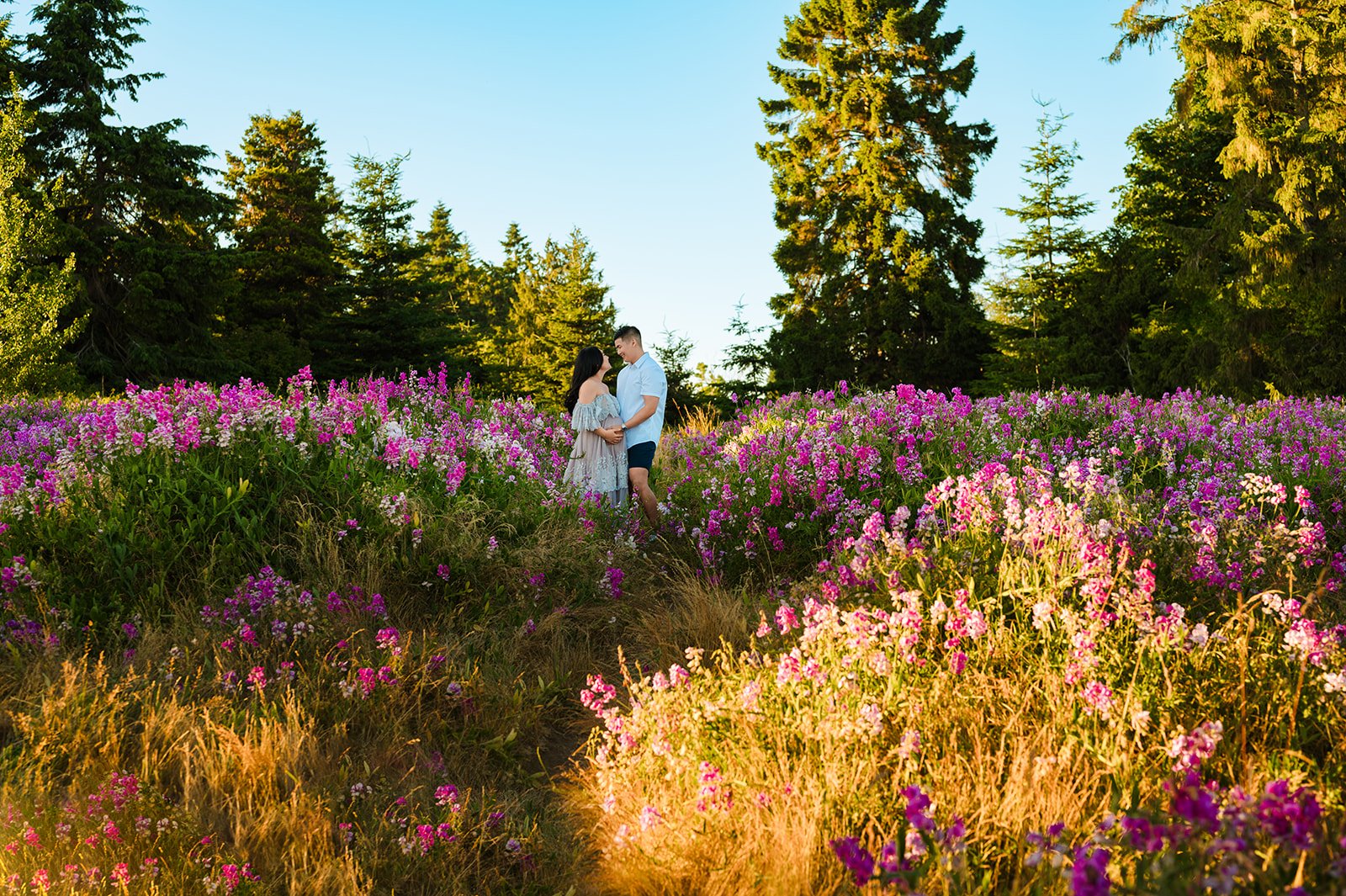 best outdoor maternity sessions in Seattle showing a husband holding wife's pregnant belly in a field of pink flowers, tall grass, and trees