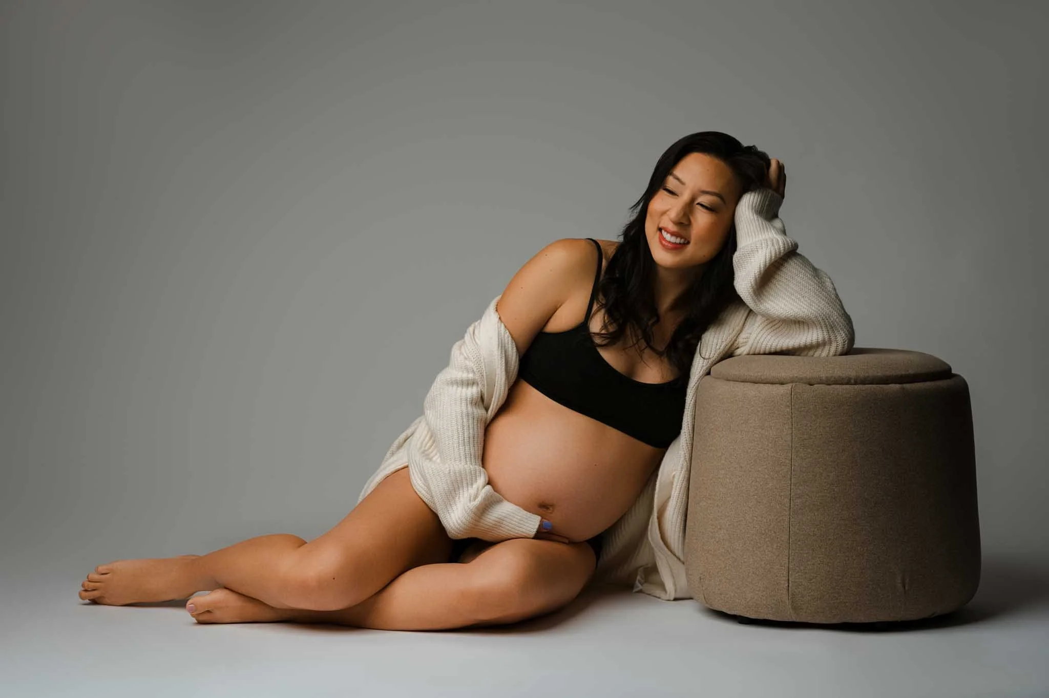 Pregnant woman wearing sports bra and cardigan sitting on floor of Seattle studio leaning on cream stool.