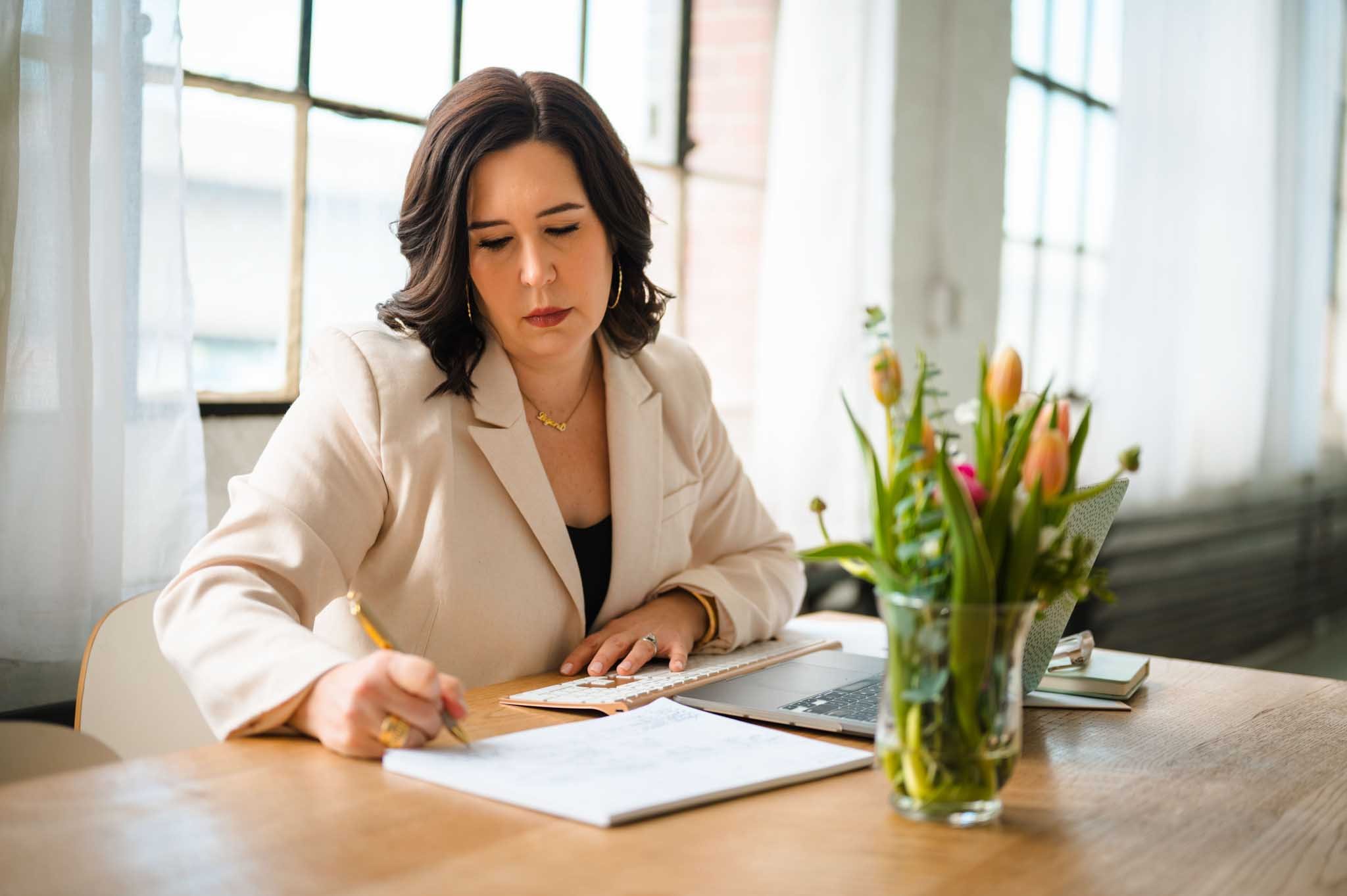 A woman with dark hair wearing a beige blazer and gold jewelry sitting at a wooden table, writing in a notebook with a pen, a laptop, and a vase with pink tulips on the table, with large windows in the background.