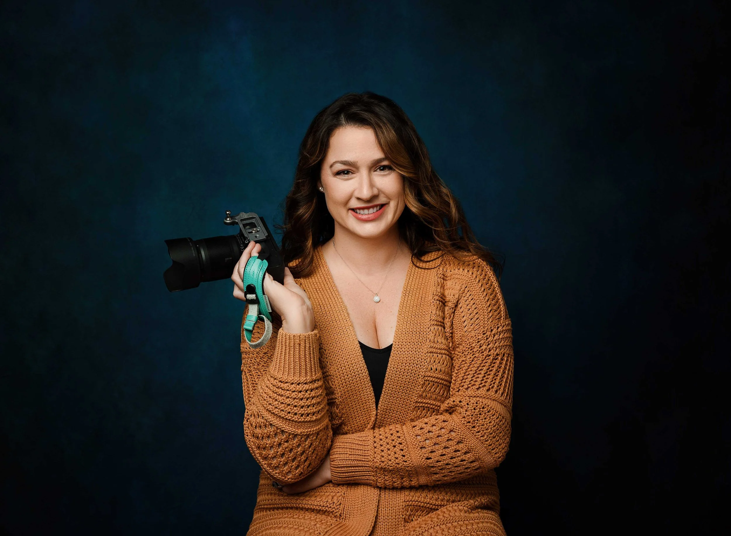 headshots near me Seattle - Smiling woman with wavy brown hair holding a professional camera with a teal strap, wearing a brown cardigan over a black top, against a dark gradient background.