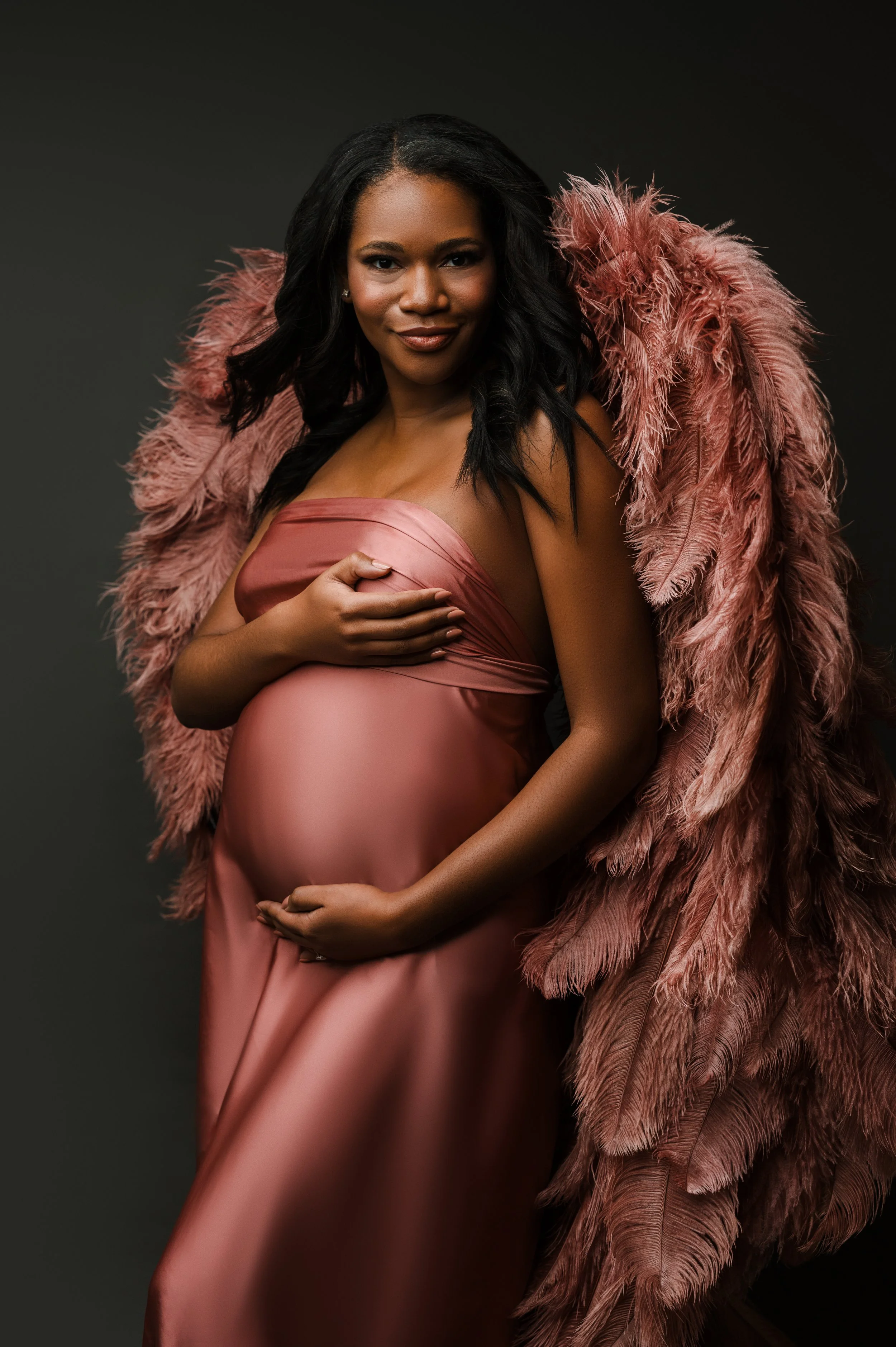 A pregnant woman in a silky pink dress, holding her belly with one hand and covering her chest with the other, wearing a large pink feathered cape, against a dark background.