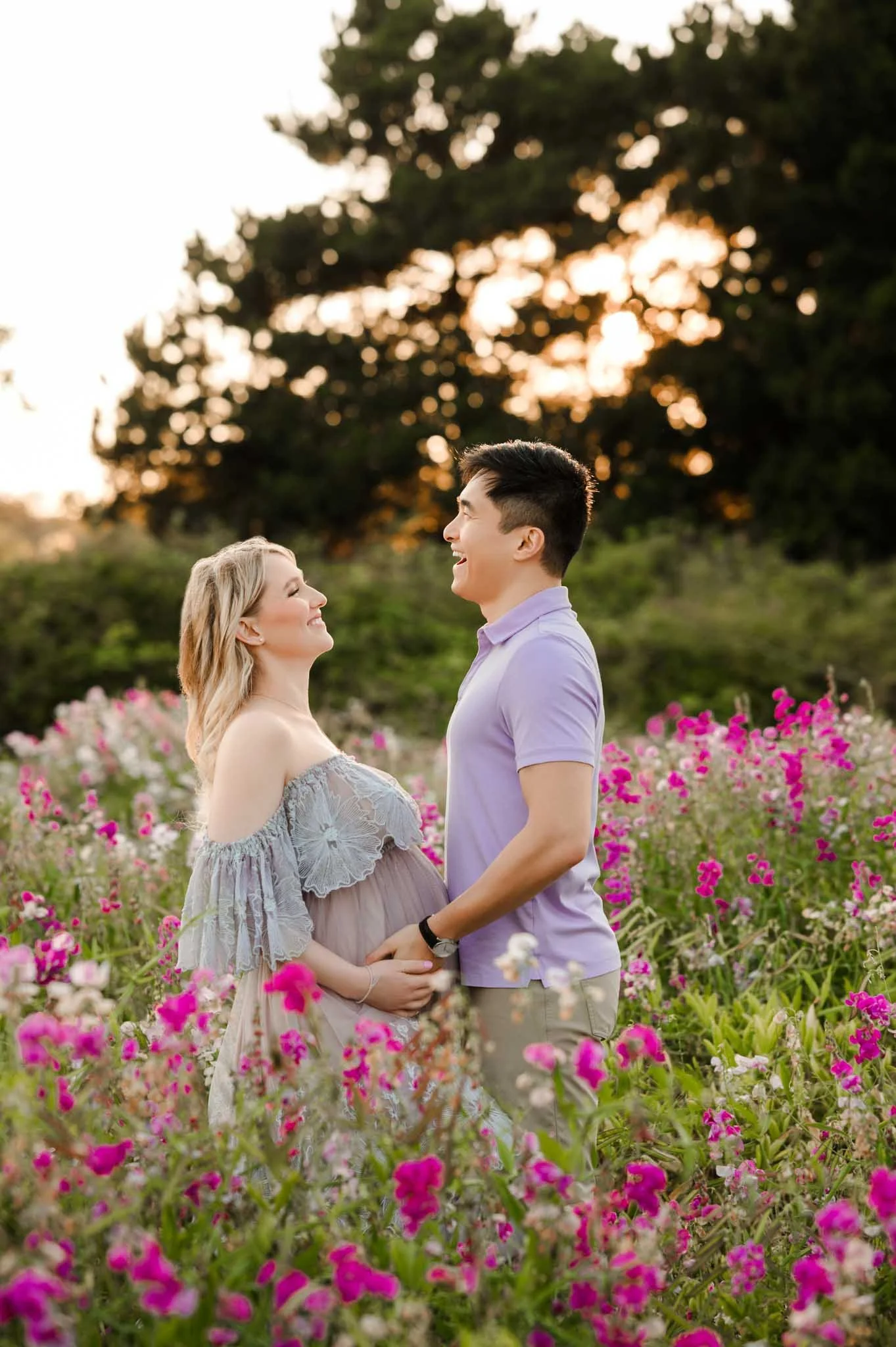 A couple standing close together in a field of pink and white flowers, smiling and looking at each other during sunset.