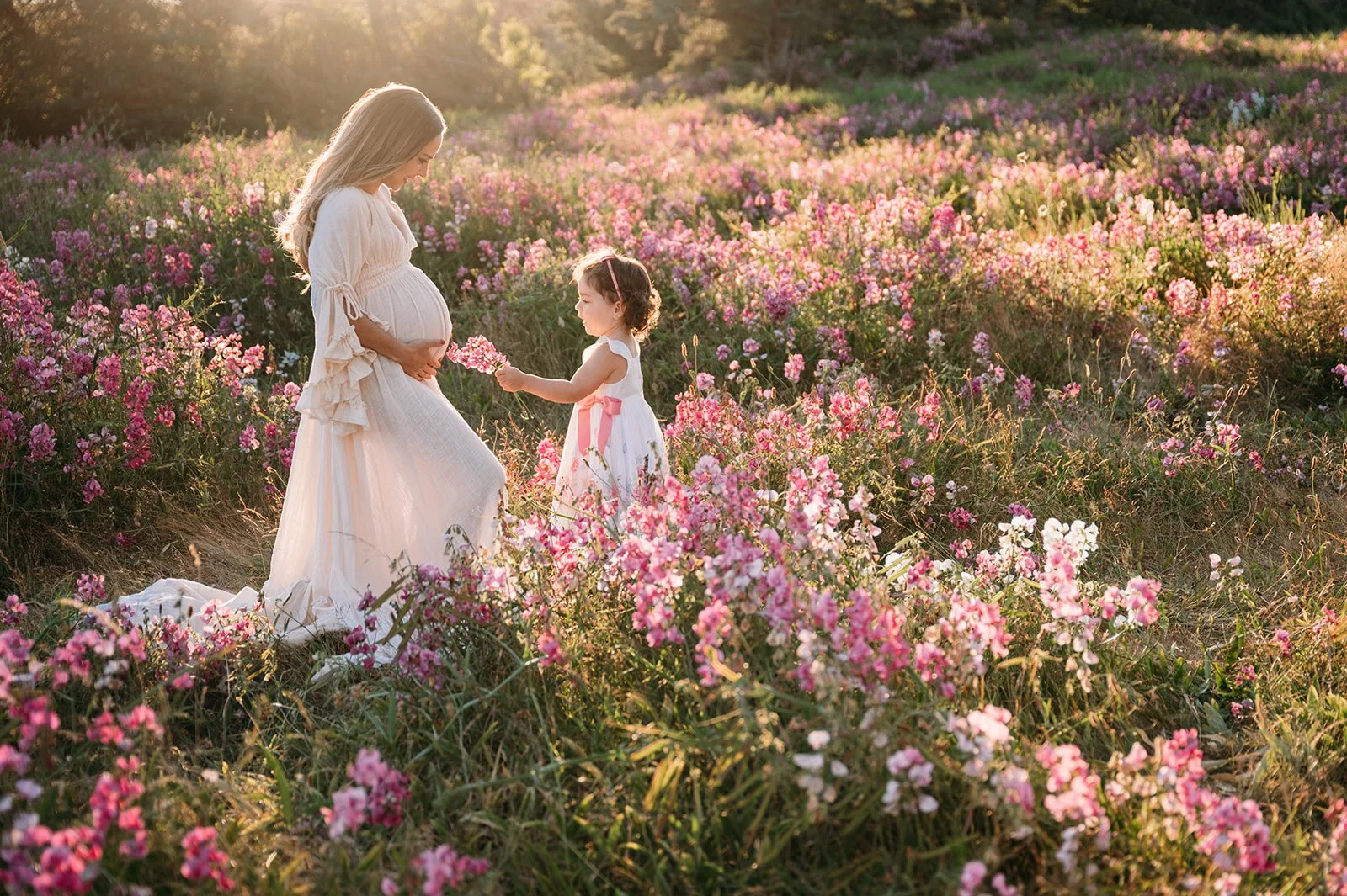 A pregnant woman and a young girl in a white dress in a field of pink and purple flowers during sunset