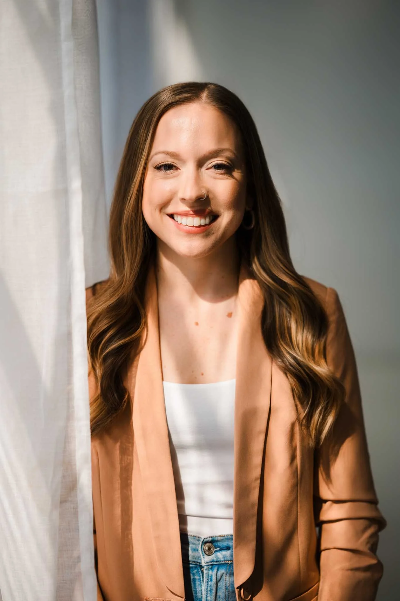 A young woman with long brown hair smiling, wearing a beige blazer and white top, standing near curtains.