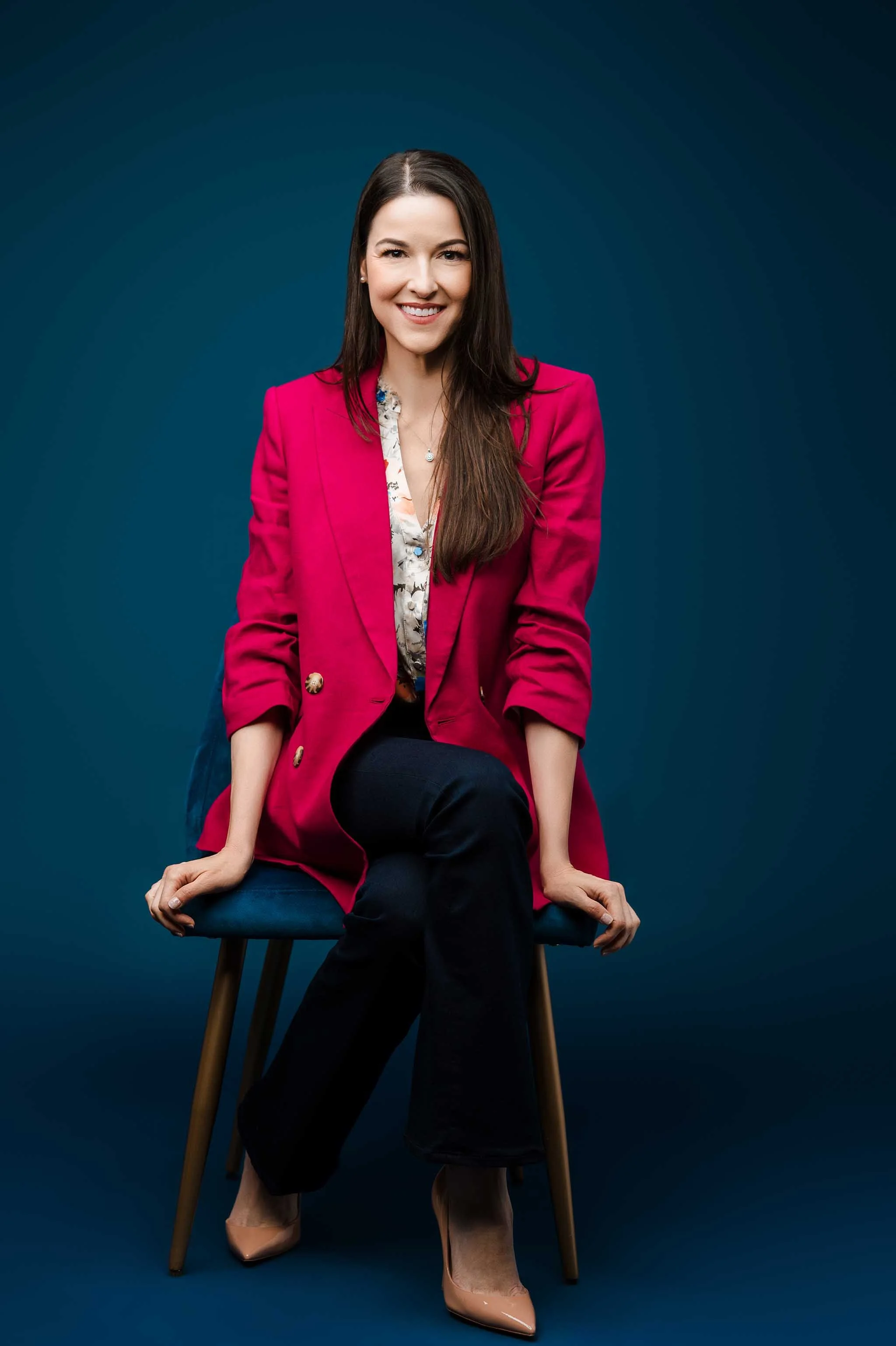 Personal branding portrait of a woman in a pink blazer seated against a blue studio background