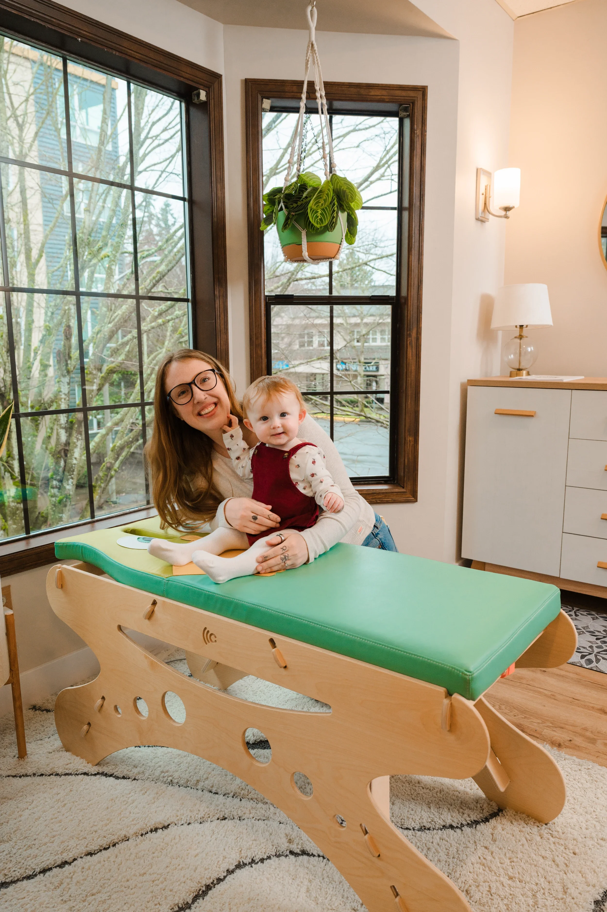 A woman with glasses and a young girl with light hair sitting on a wooden and green medical examination table indoors, smiling at the camera. The room has large windows with views of trees outside and is decorated with modern furniture and a hanging plant.